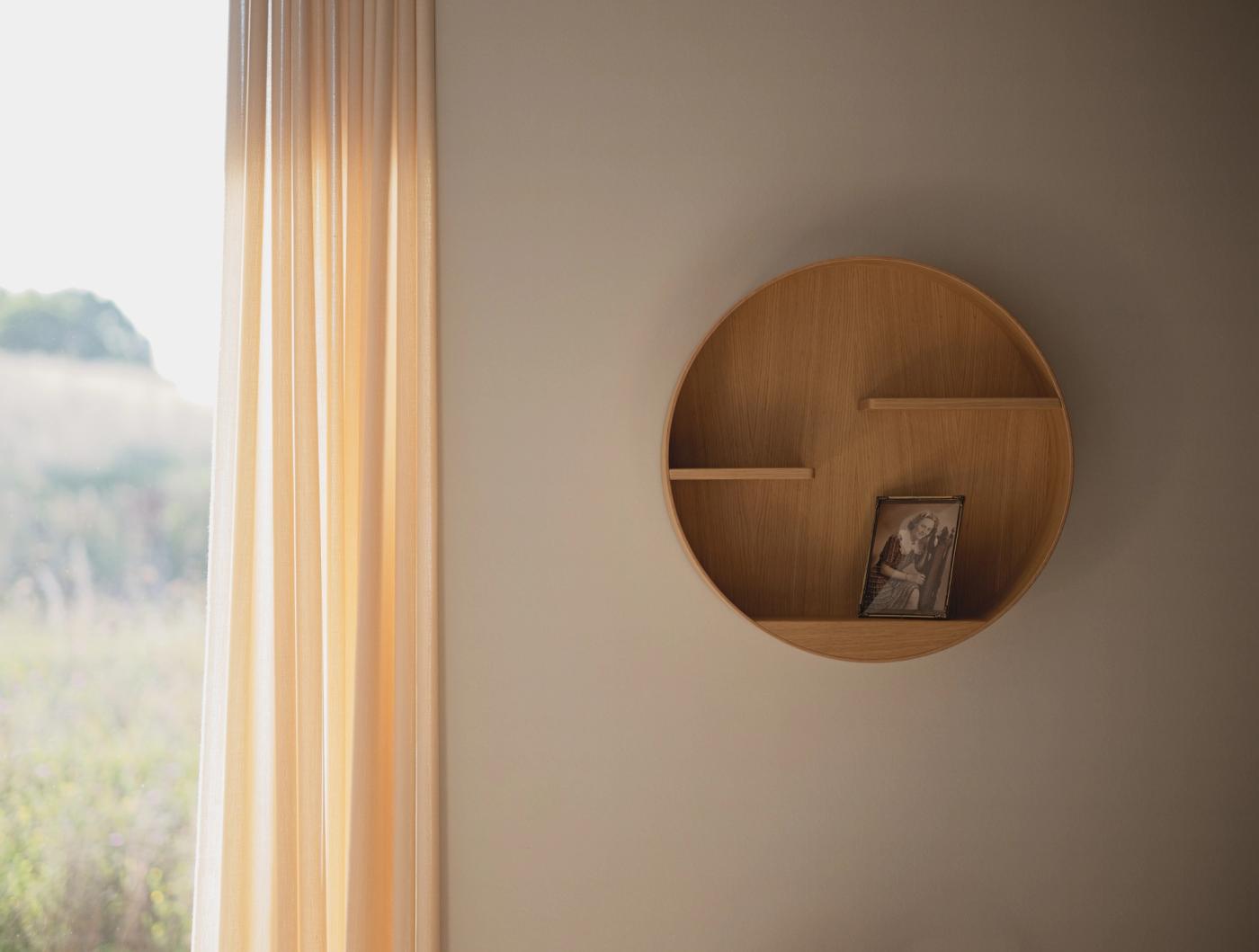 Cyklus memorial shelf in oak with brass details, a round shelf holding a small framed photograph, on a wall beside sheer curtains and a window.