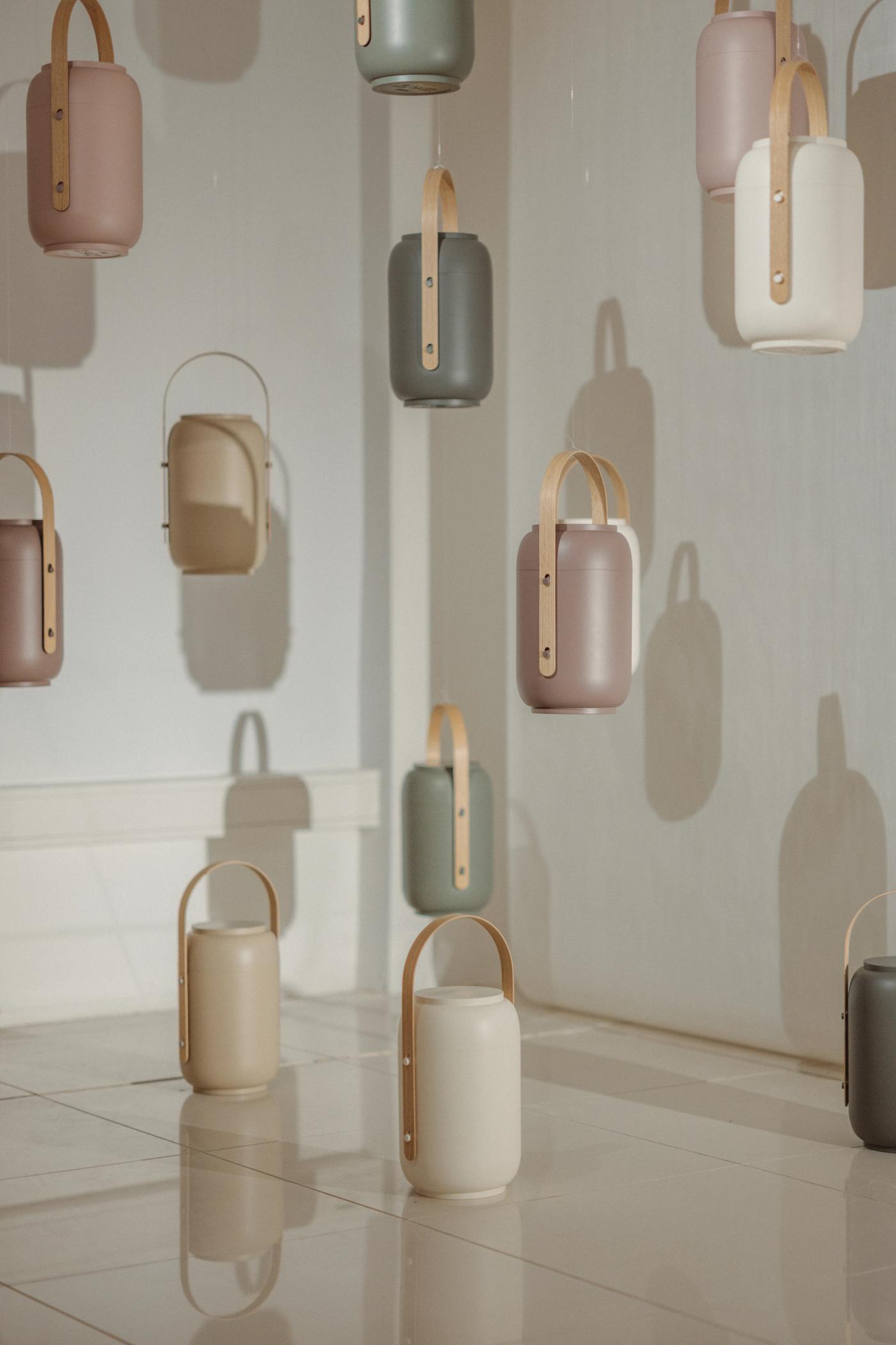 Akari urns in sage, rose, cream, taupe and grey with wooden handles, hanging on a white wall and standing on a tiled floor, beside an oak armchair.