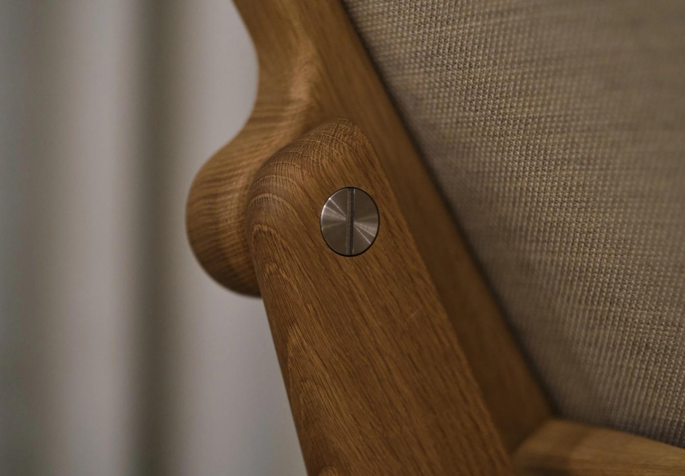 Close-up of an oak armrest on a GAZE chair with a round stainless steel fitting, beside light upholstery.