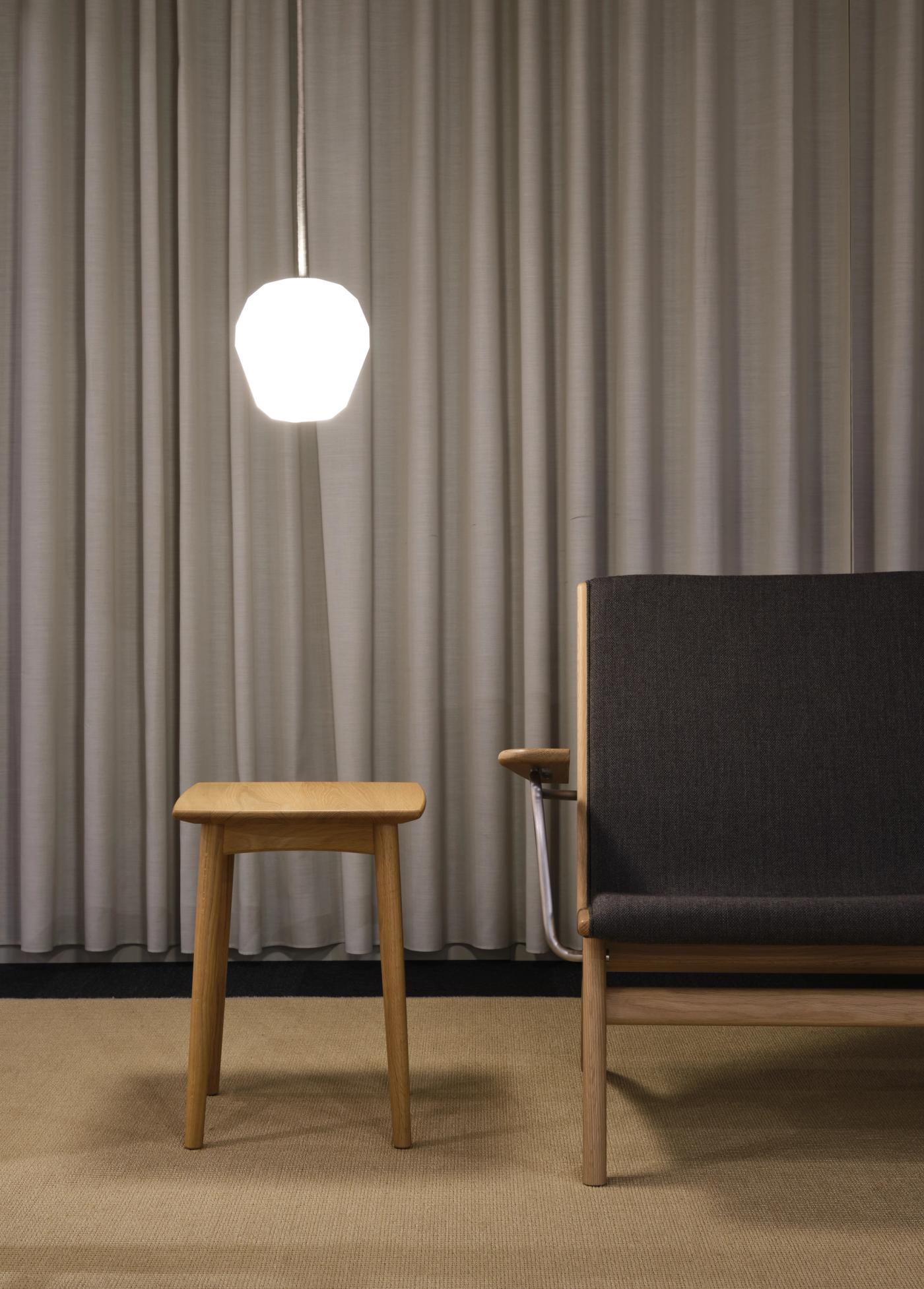 GAZE lounge chair with a dark grey upholstered seat and oak frame beside a small oak side table, under a white pendant lamp and grey curtains.