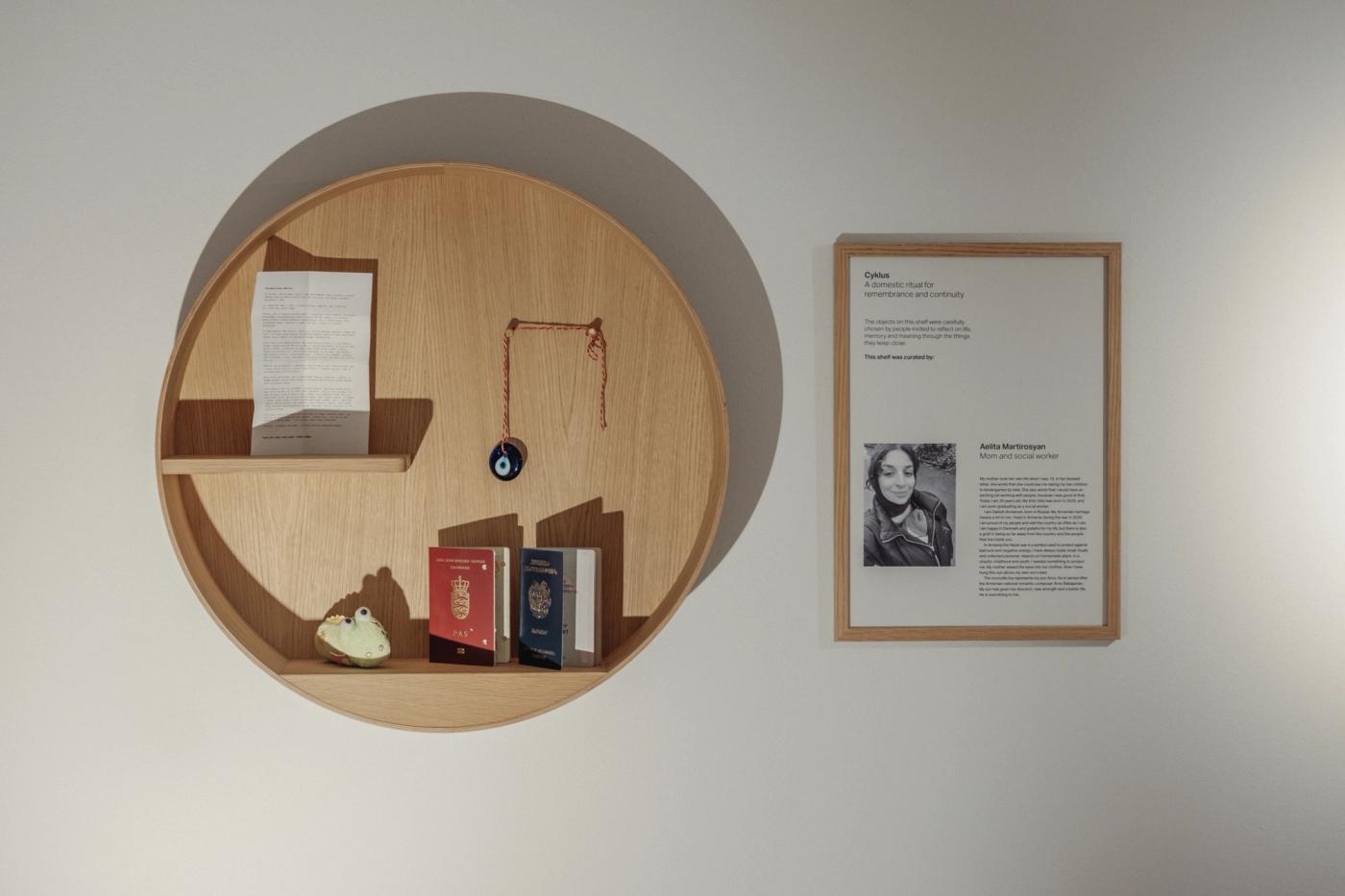 Alt-tekst:  Cyklus memorial shelf in oak holding a folded paper, a small charm on a chain, two passports and a small ceramic object, beside a framed portrait and text panel.