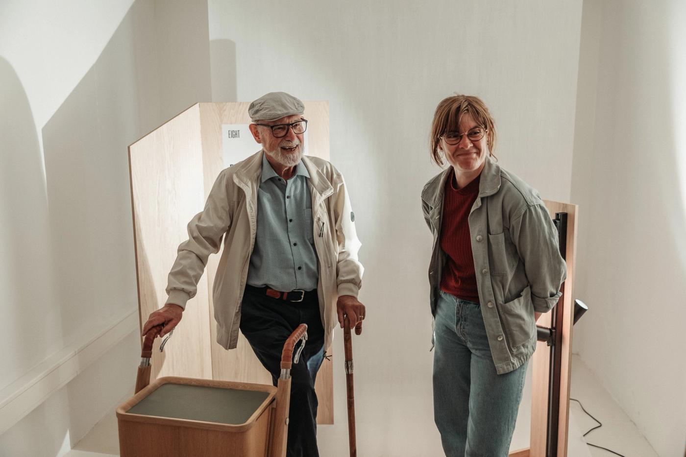 Older man in a flat cap and glasses leaning on a Sindig wooden walker with a grey seat panel, beside a smiling woman in a grey jacket, in a bright white room.