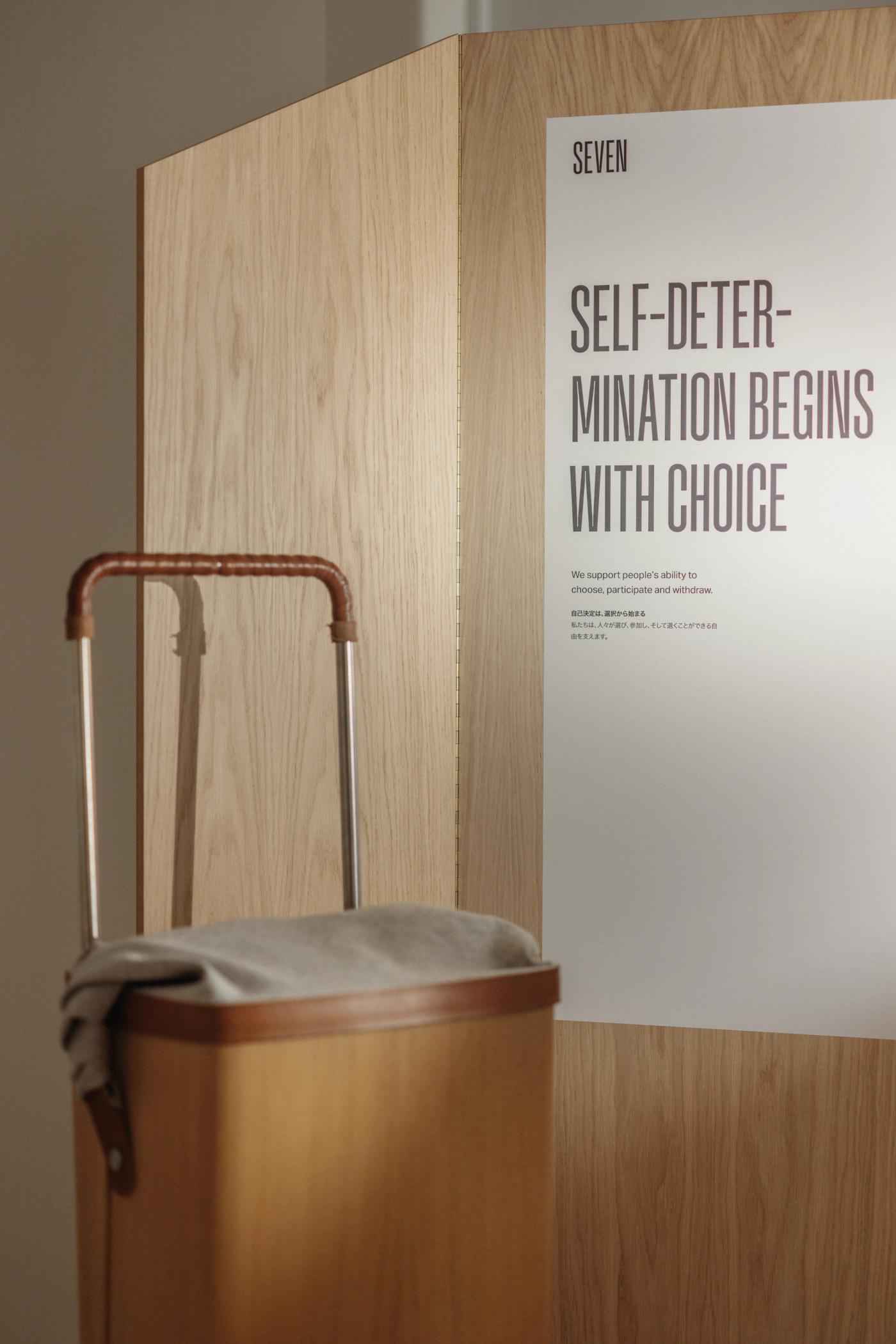 Raku trolley in oak with a wool-lined basket and a leather-wrapped steel handle, in front of an oak panel with the poster "Self-determination begins with choice".