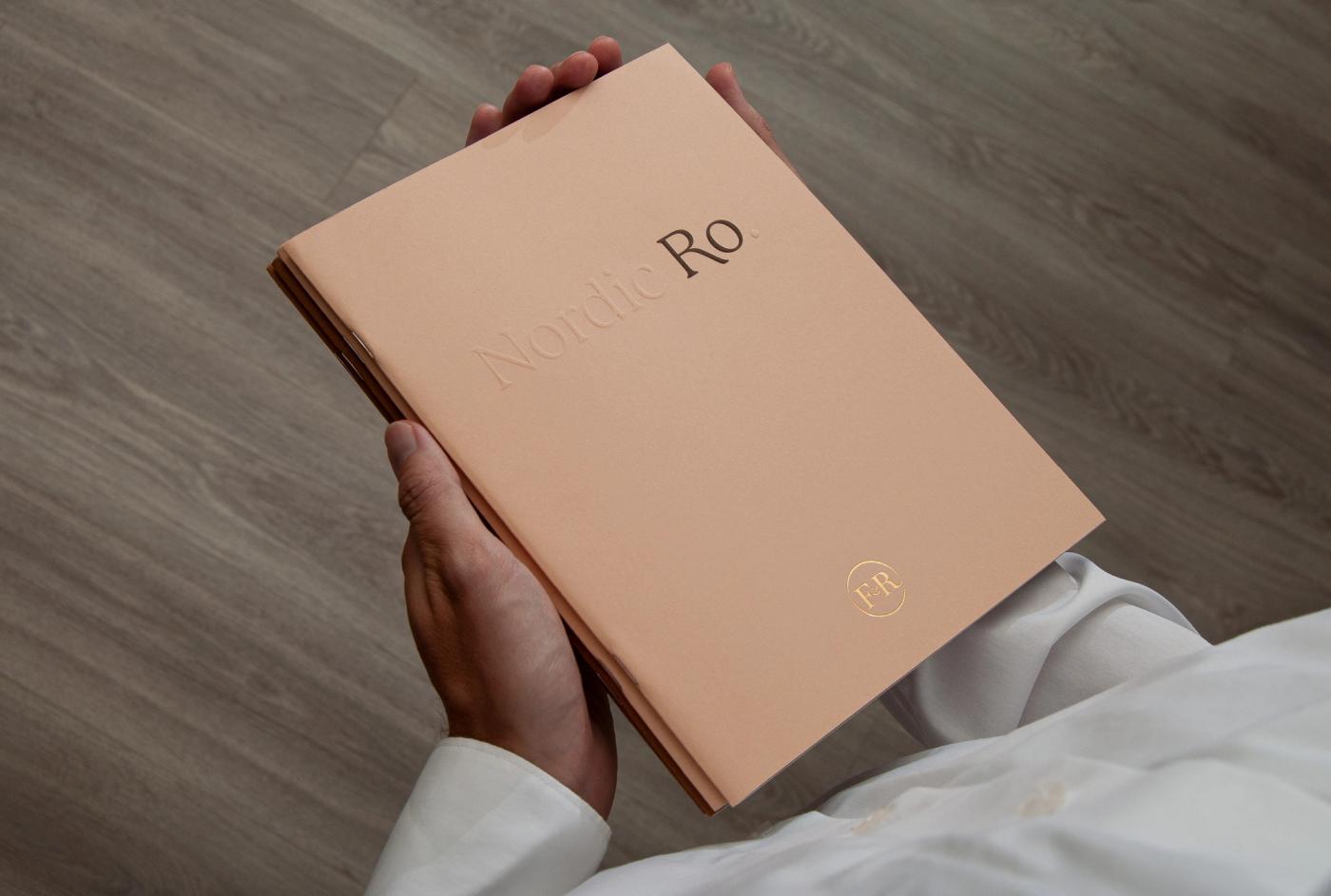 Hands holding the Ro catalogue with a peach cover reading "Nordic Ro" and a gold logo, over a wooden floor.