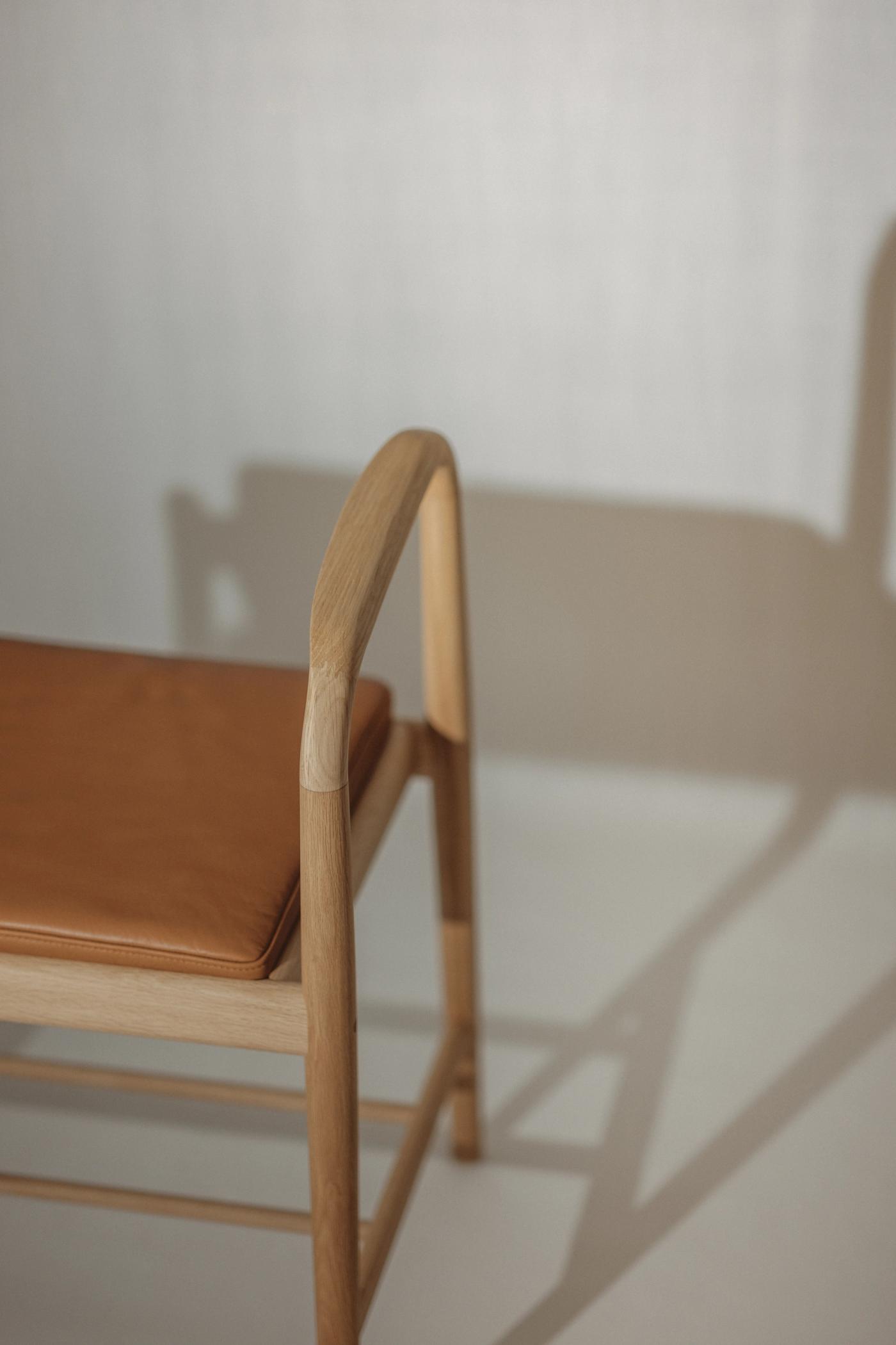 Oak chair with a curved armrest and a tan leather seat cushion, shown from the side against a pale wall with a soft shadow.