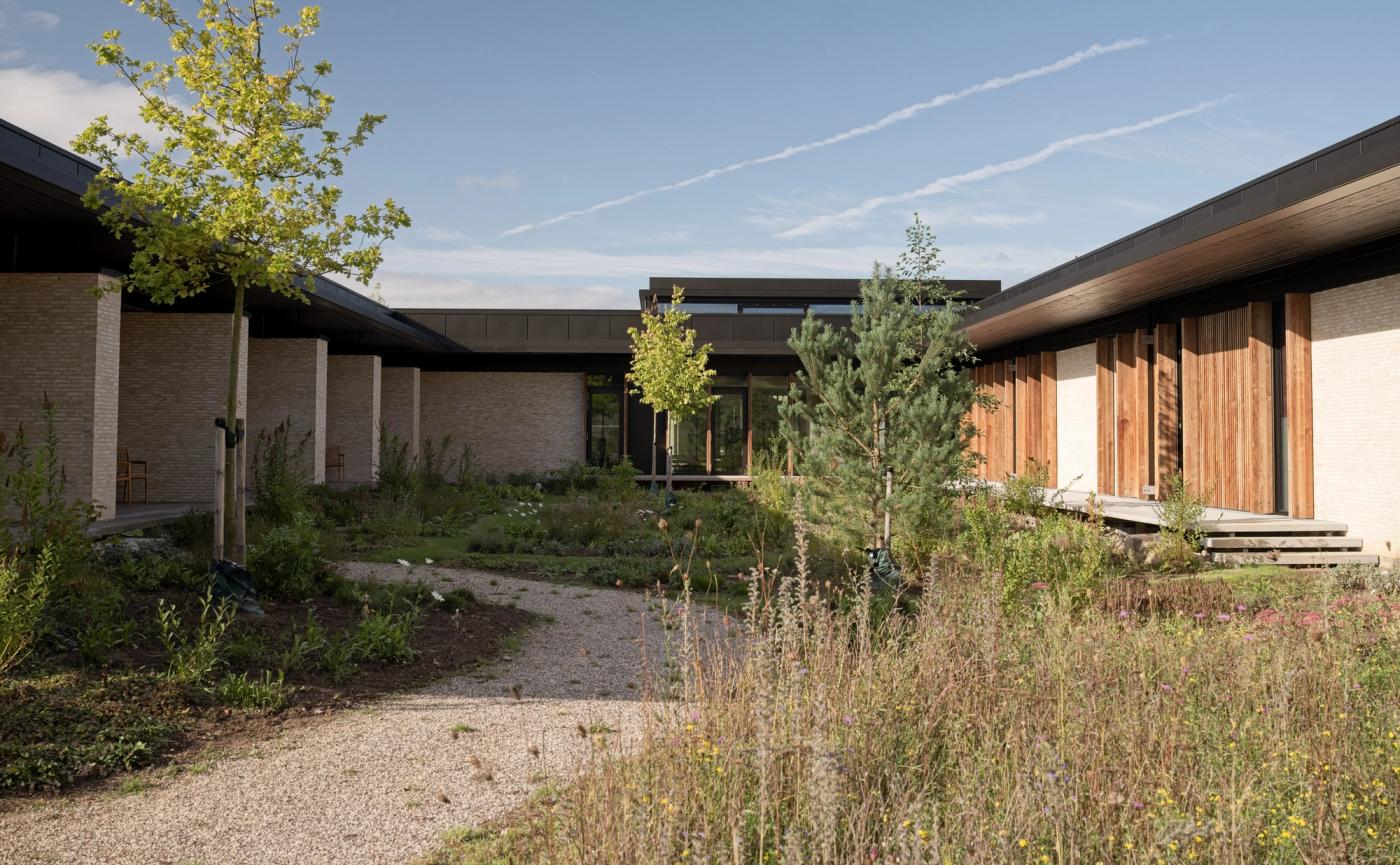 Exterior of the Privathospitalet Mølholm building in Vejle, with light brick walls, timber-clad doors, a gravel path and a garden of trees and wild grasses under a blue sky.