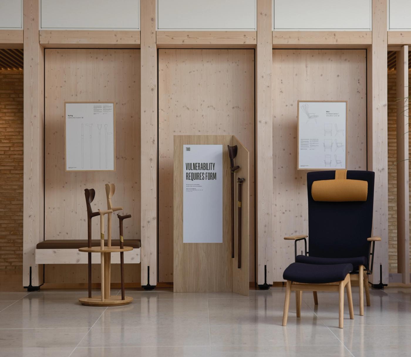 Wide view of an exhibition space with light wood walls, a walking stick stand and a navy lounge chair with an ochre headrest, beside a panel with the poster "Vulnerability Requires Form".