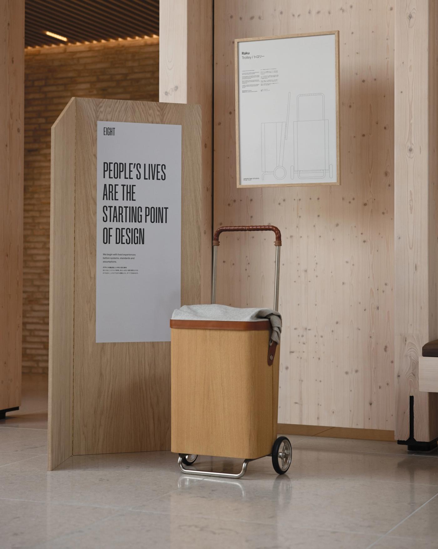 Raku trolley in oak with a wool-lined bag, leather-wrapped handle and stainless steel frame, standing beside a wooden panel with the poster "People's Lives Are the Starting Point of Design".