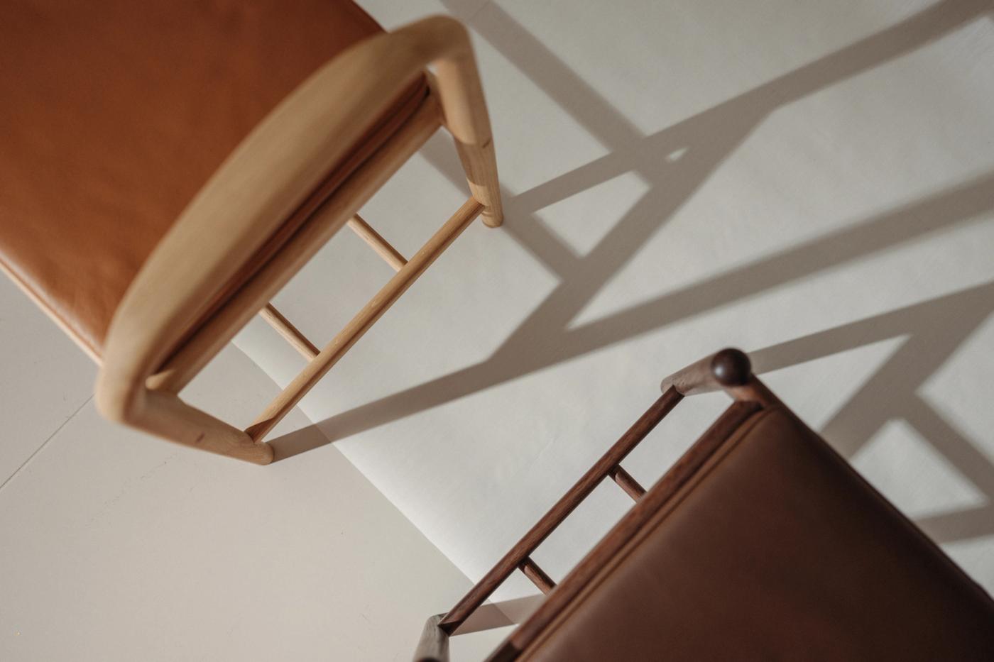 Overhead view of two chairs with tan leather seats and light wooden frames, casting long diagonal shadows on a pale wall.