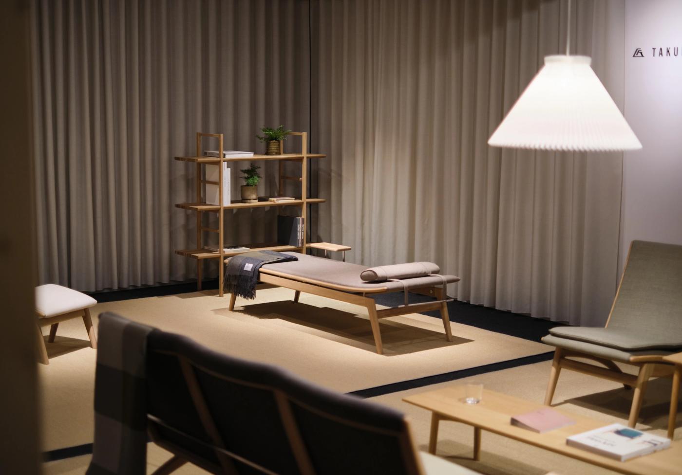 GAZE daybed in oak with a grey upholstered seat and cushion, placed beside an oak shelving unit and a low oak table, under a white pendant lamp in a room with grey curtains.