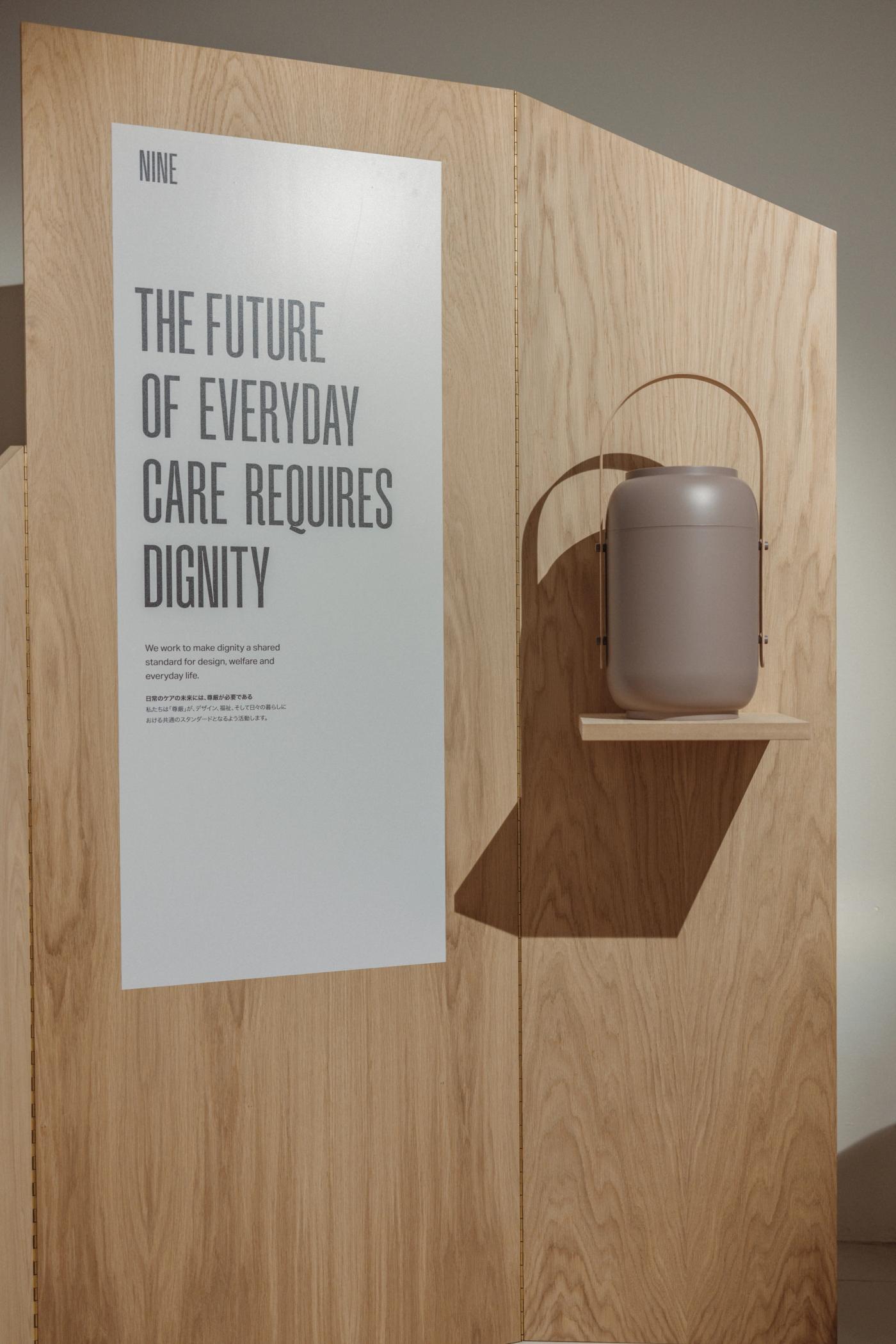 Oak wall panel with the poster "The future of everyday care requires dignity" and an Akari urn in taupe with a wooden handle, hanging on a small oak shelf.