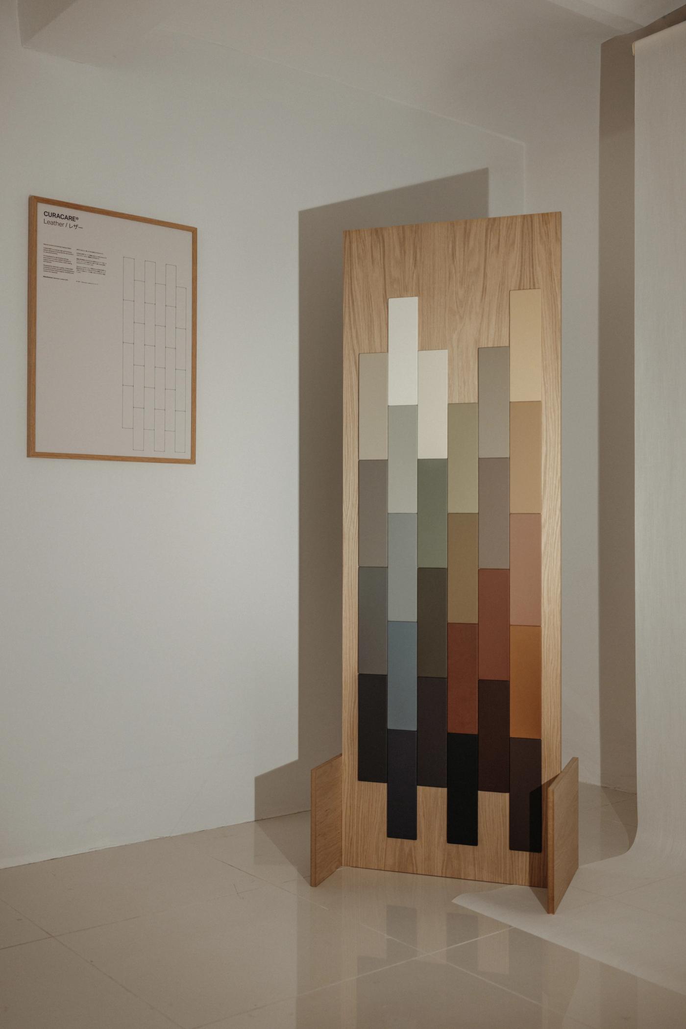 Oak sample panel with rows of colour and material swatches in cream, grey, beige, rust and dark brown, standing beside a framed technical drawing on a white wall.