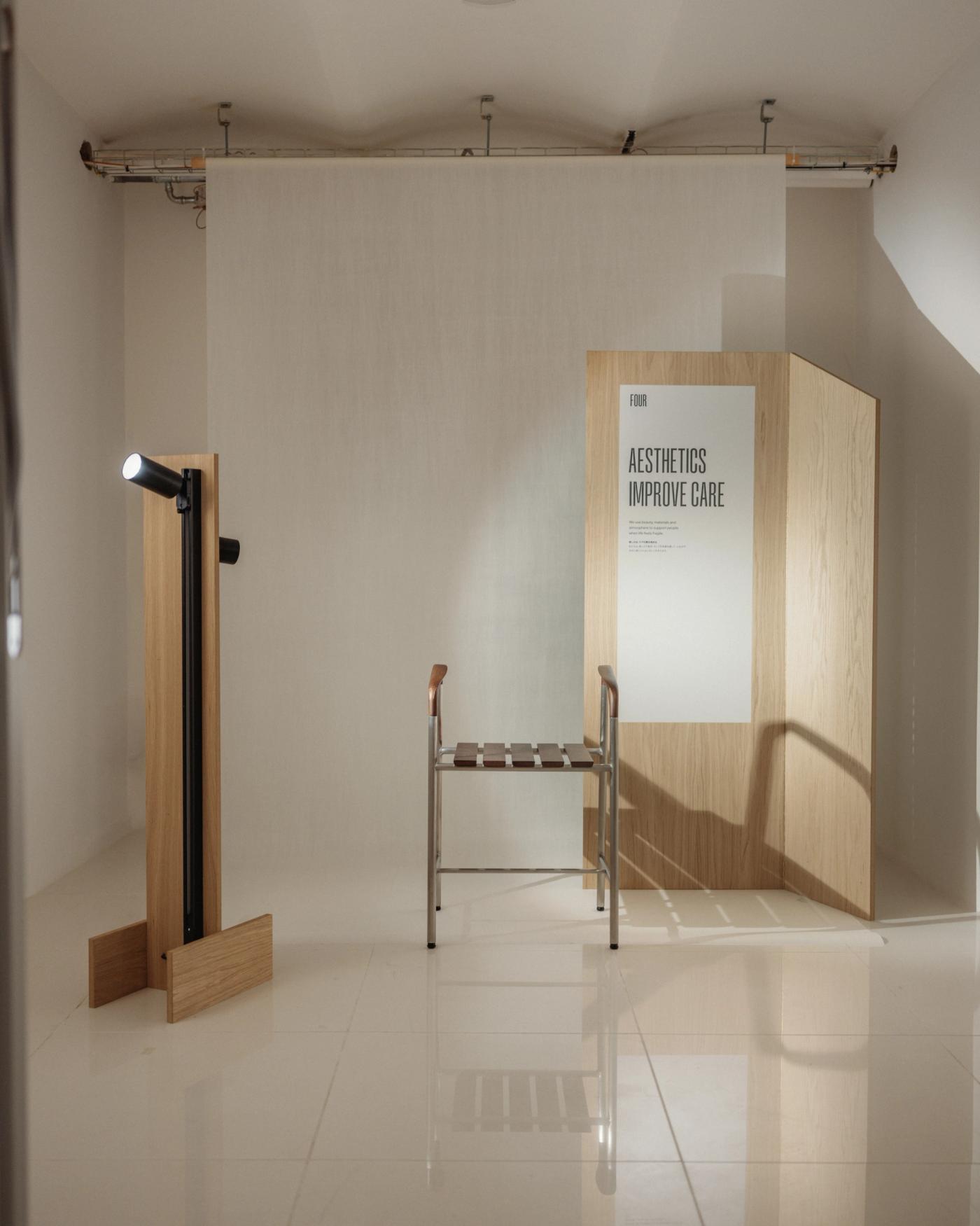 Exhibition room with a bath stool in stainless steel and wood in the centre, beside an oak wall lamp on the left and an oak panel with the poster "Aesthetics Improve Care" on the right.