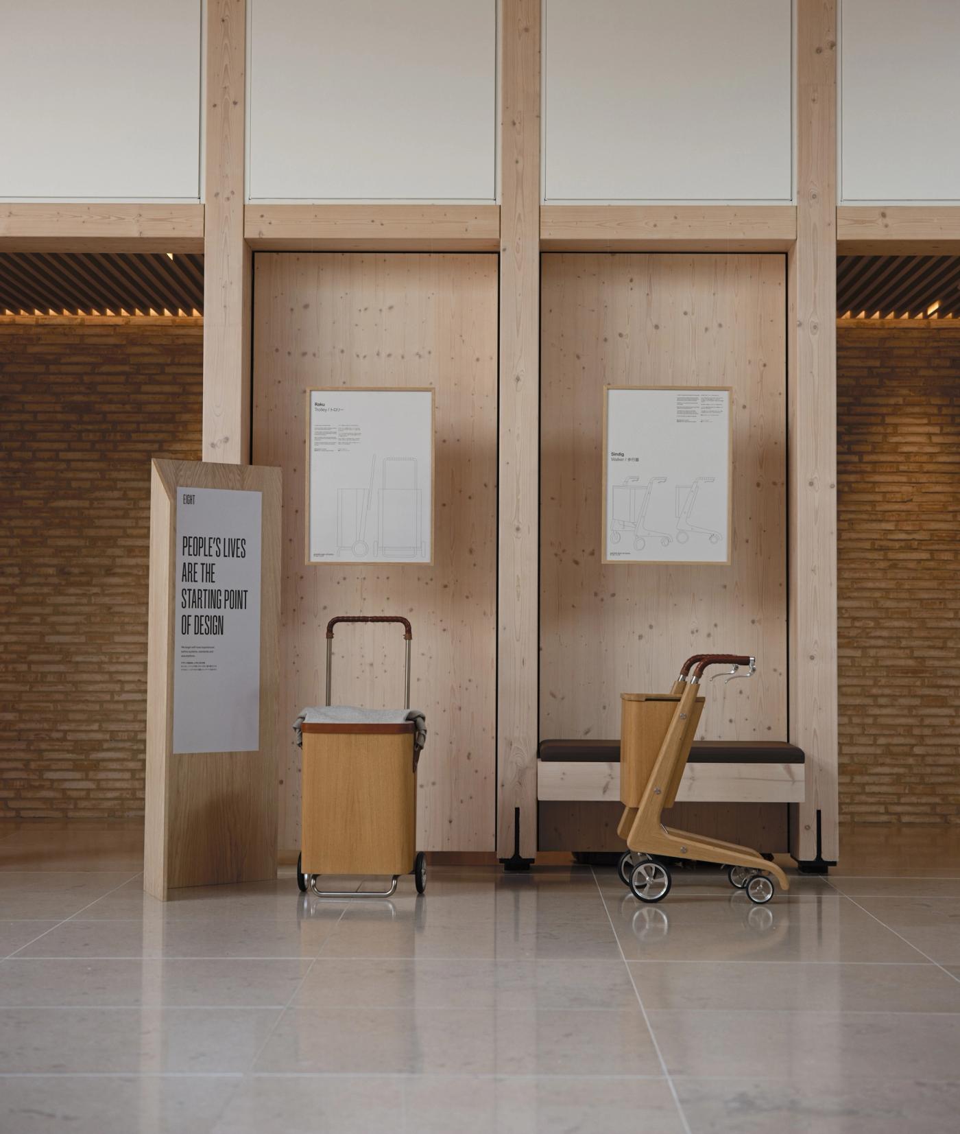 Raku trolley in oak and Sindig wooden walker with wheels, standing side by side in an exhibition space with a bench and a wooden panel with the poster "People's Lives Are the Starting Point of Design".