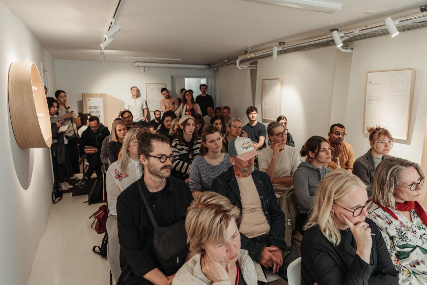 Audience seated and standing in a white gallery room during a talk at The Design We Hide Away, with framed drawings on the wall and a round oak shelf on the left.