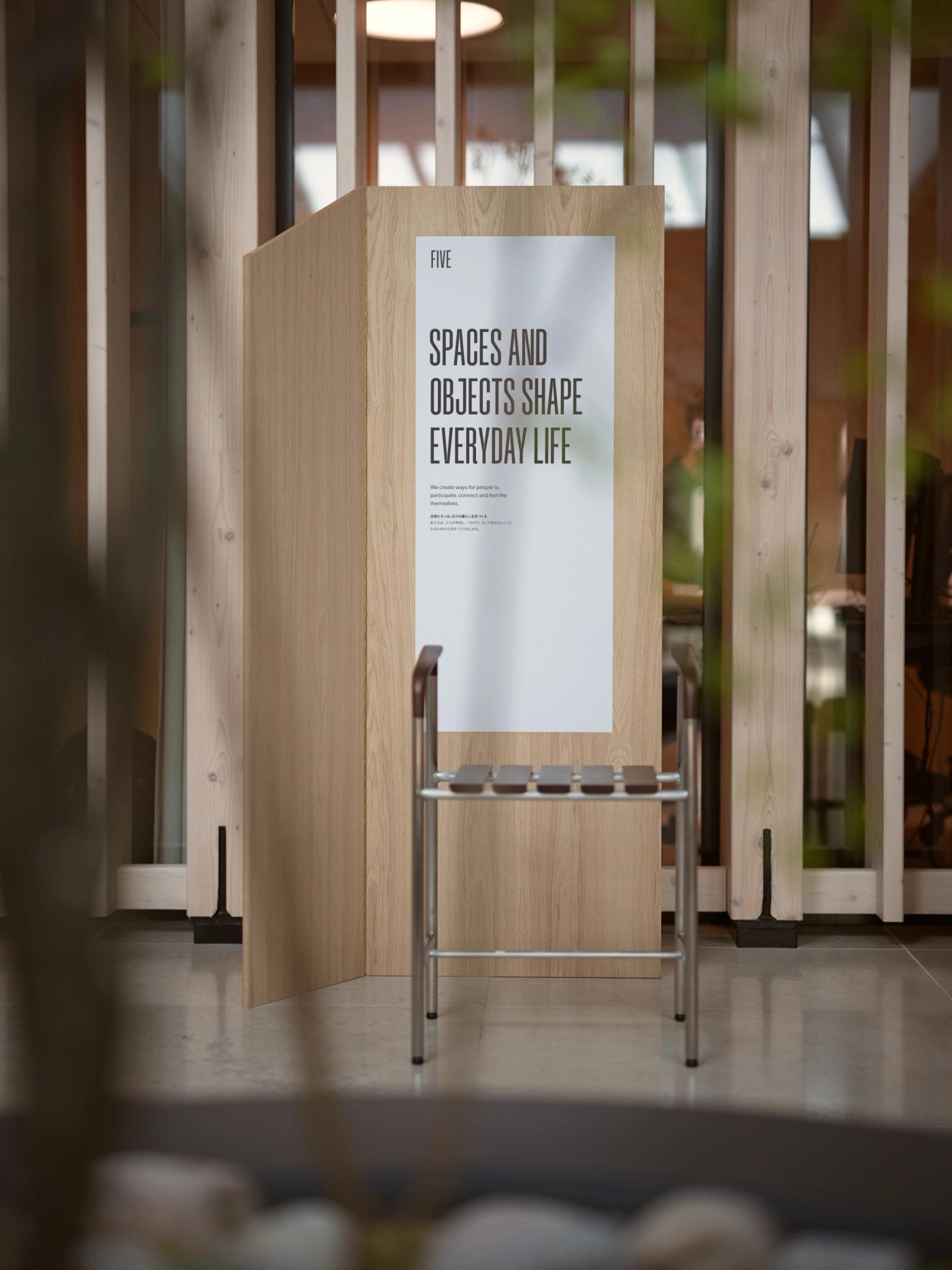Bath stool in wood and stainless steel with a slatted seat, placed beside a wooden panel with the poster "Spaces and Objects Shape Everyday Life".