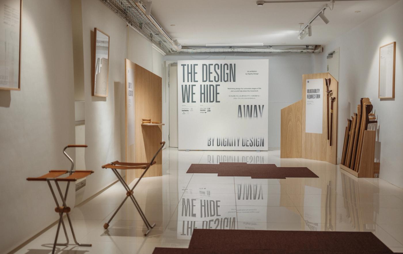 Entrance to The Design We Hide Away exhibition with the large poster "The Design We Hide Away by Dignity Design" at the end of a white gallery, a folding wooden stool with a leather seat on the left and a stand of wooden canes on the right.