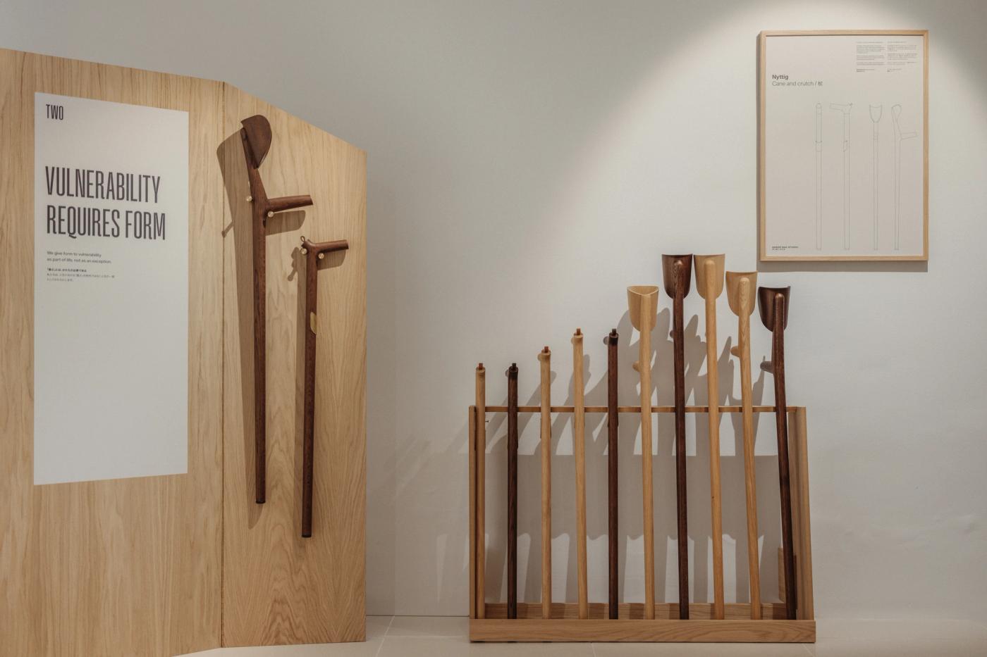 Wooden crutches and walking sticks in light and dark wood, hung on an oak panel with the poster "Vulnerability requires form" and displayed in a rack of canes of different heights, beside a framed drawing on the white wall.
