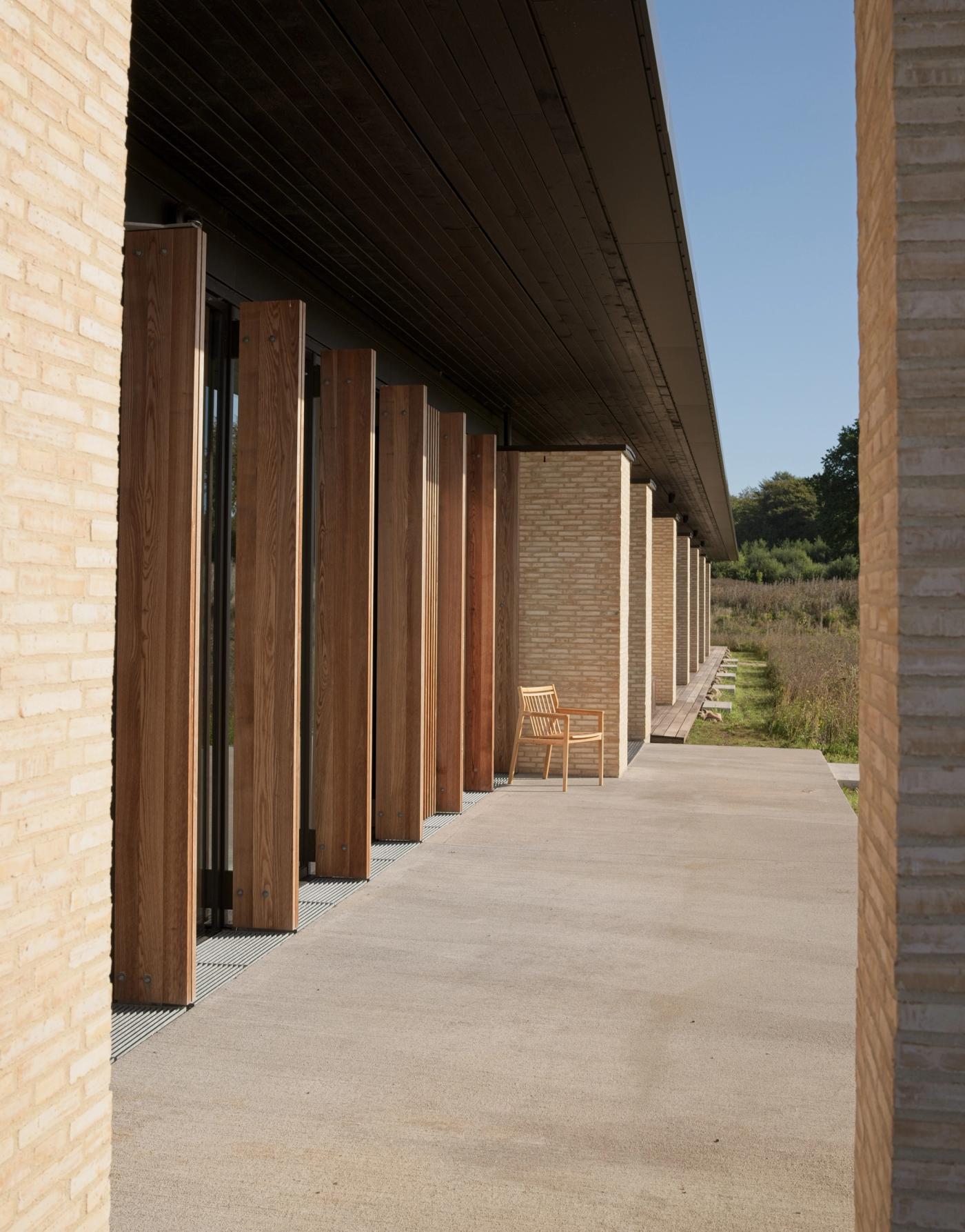 Covered walkway at the Privathospitalet Mølholm building, with light brick pillars, timber-slatted walls, a concrete terrace and a wooden chair facing the garden.