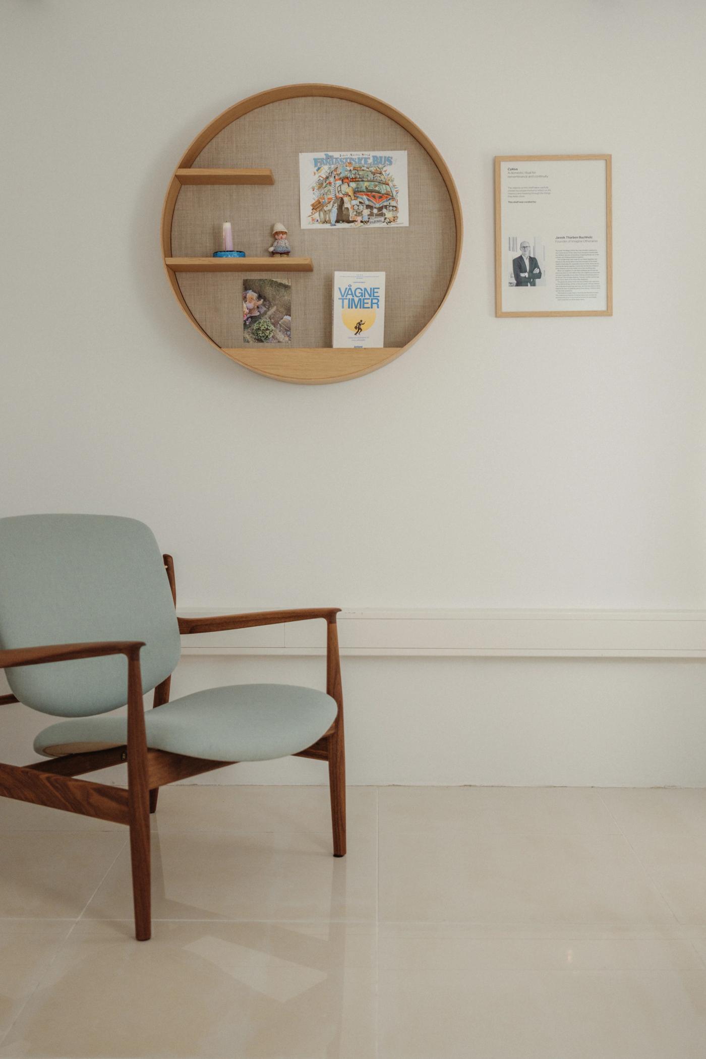 Cyklus memorial shelf in oak with a woven backing, holding a few small objects and printed matter, beside a framed text panel and a wooden lounge chair with a mint upholstered seat