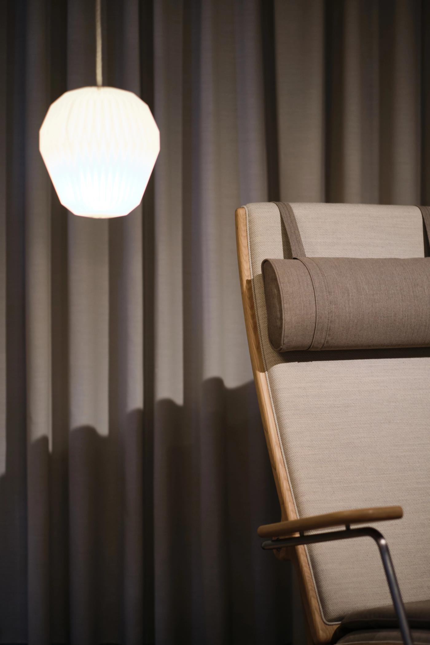Close-up of a GAZE high-back chair with a light upholstered back, a neck cushion and an oak armrest with a steel support, beside grey curtains and a white pendant lamp.