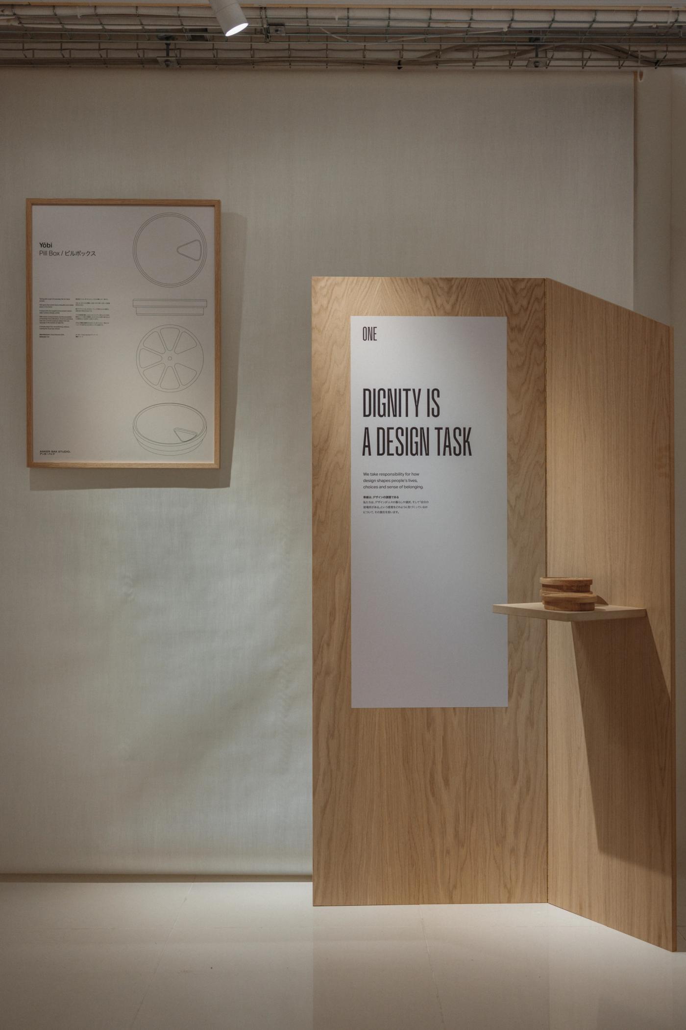 Oak wall panel with the poster "Dignity is a design task" and a small wooden bowl on a shelf, beside a framed technical drawing of circular forms on a pale wall.