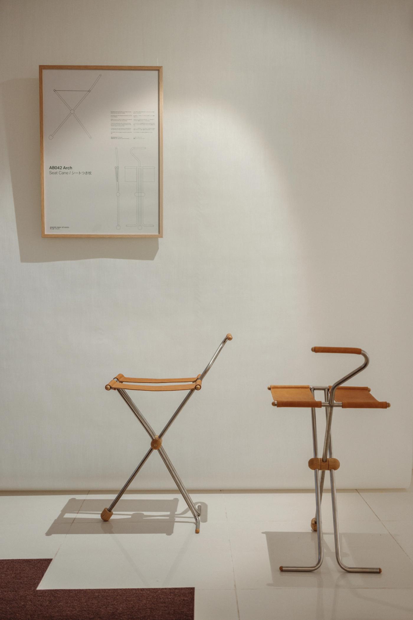 Folding stool with a leather seat and a steel X-frame, beside a seat cane with a leather seat, a curved handle and a steel frame, in front of a white wall with a framed drawing.