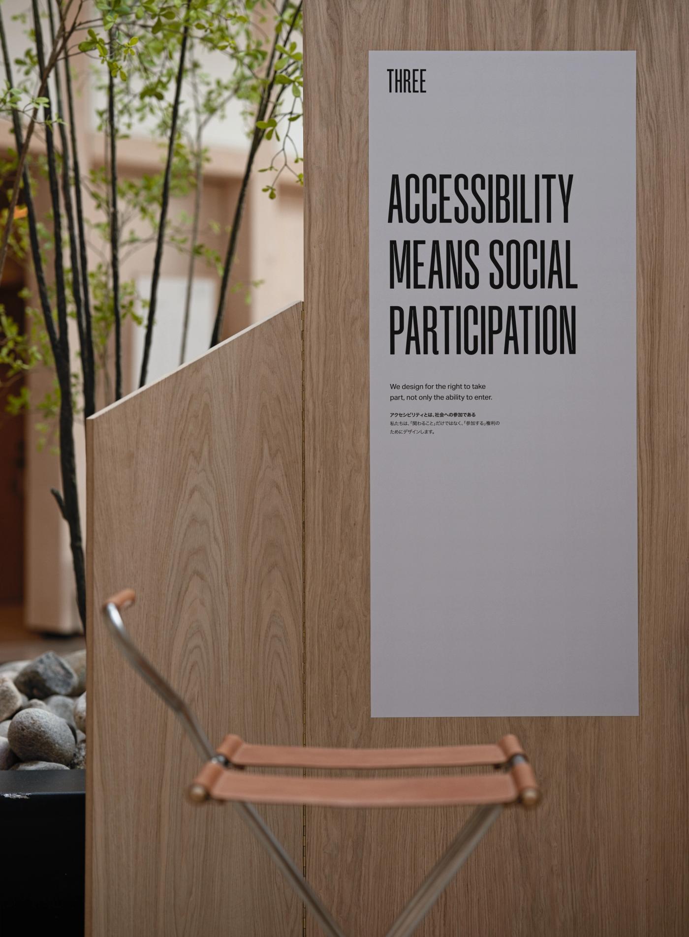 Folding stool with a tan leather seat and steel legs in front of a wooden panel with the poster "Accessibility Means Social Participation", with a plant and stones on the left.
