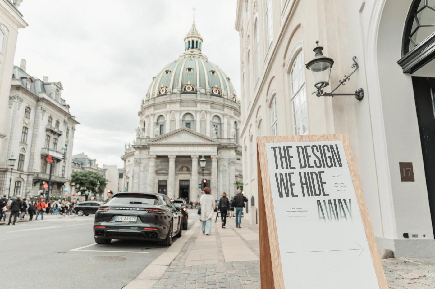 Street sign for The Design We Hide Away on a wooden stand outside a white building in Copenhagen, with the domed Marble Church and a dark car on the street behind it.