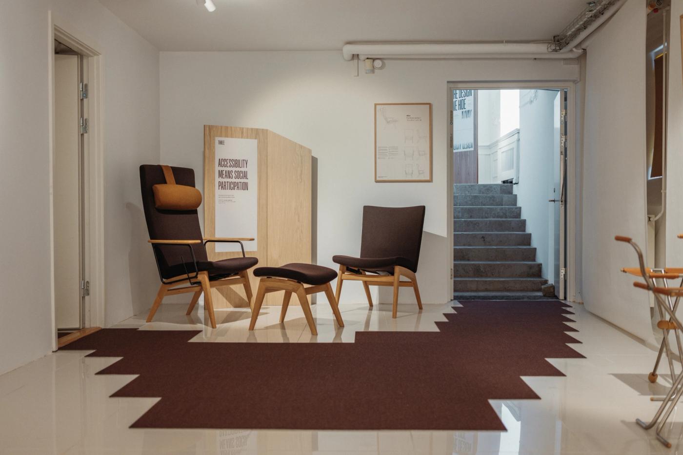 Hiro recliner chair with a dark brown upholstered seat, a tan headrest and an oak frame, beside a matching footstool and a second brown armchair on a brown rug, with the poster "Accessibility means social participation" on an oak panel behind them.