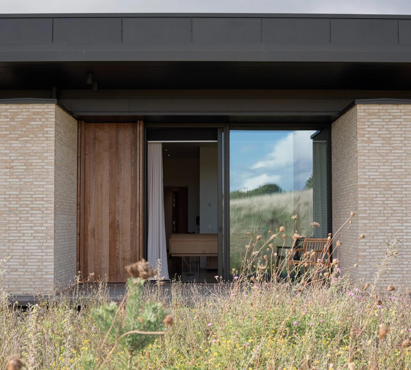 Exterior of a Nordic house with light brick walls, a wooden-framed glass door and a view into the interior, with a meadow of wild grasses in front.