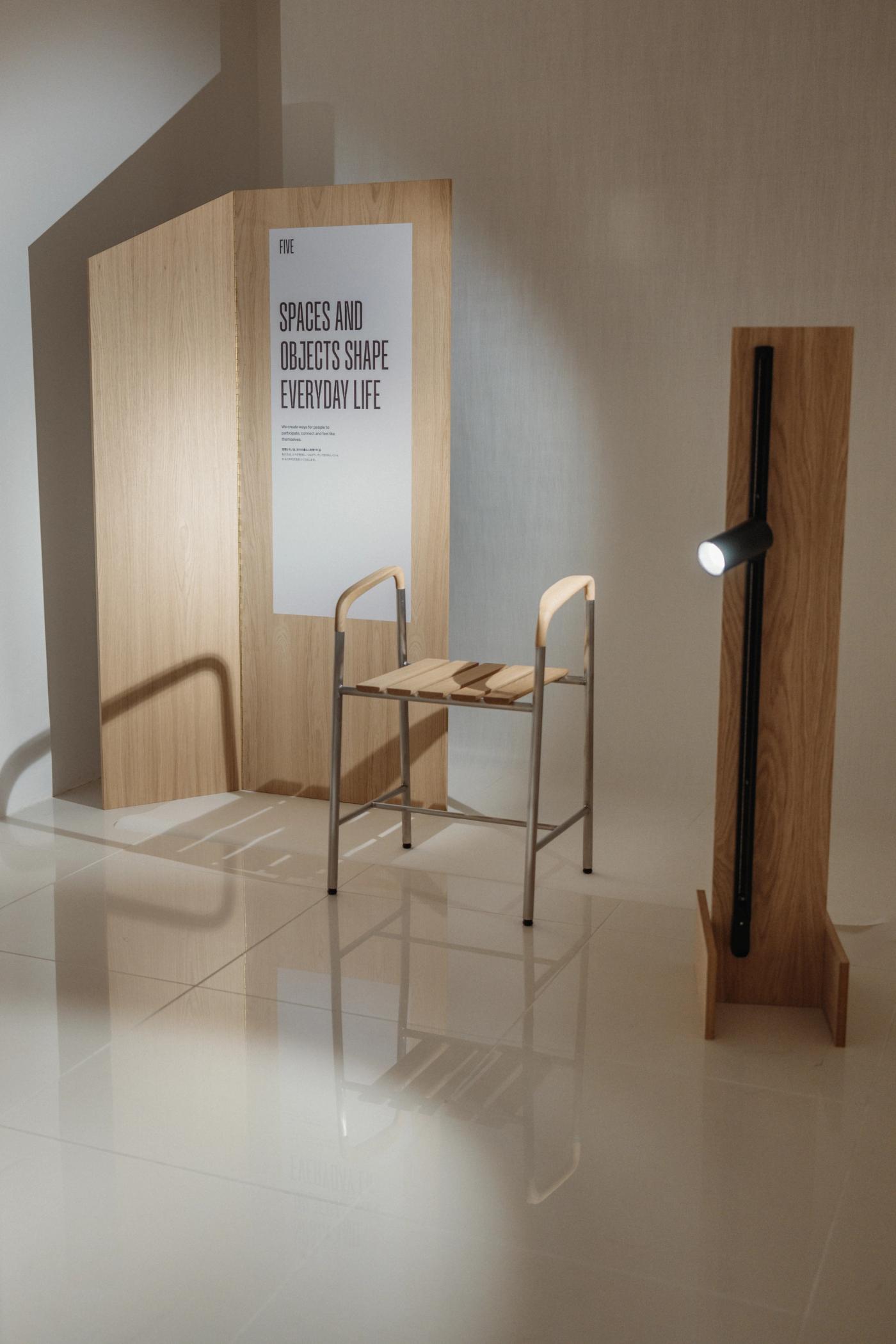 Steel-framed chair with oak armrests and a slatted oak seat beside a tall oak wall lamp with a black spotlight, in front of an oak panel with the poster "Spaces and Objects Shape Everyday Life".