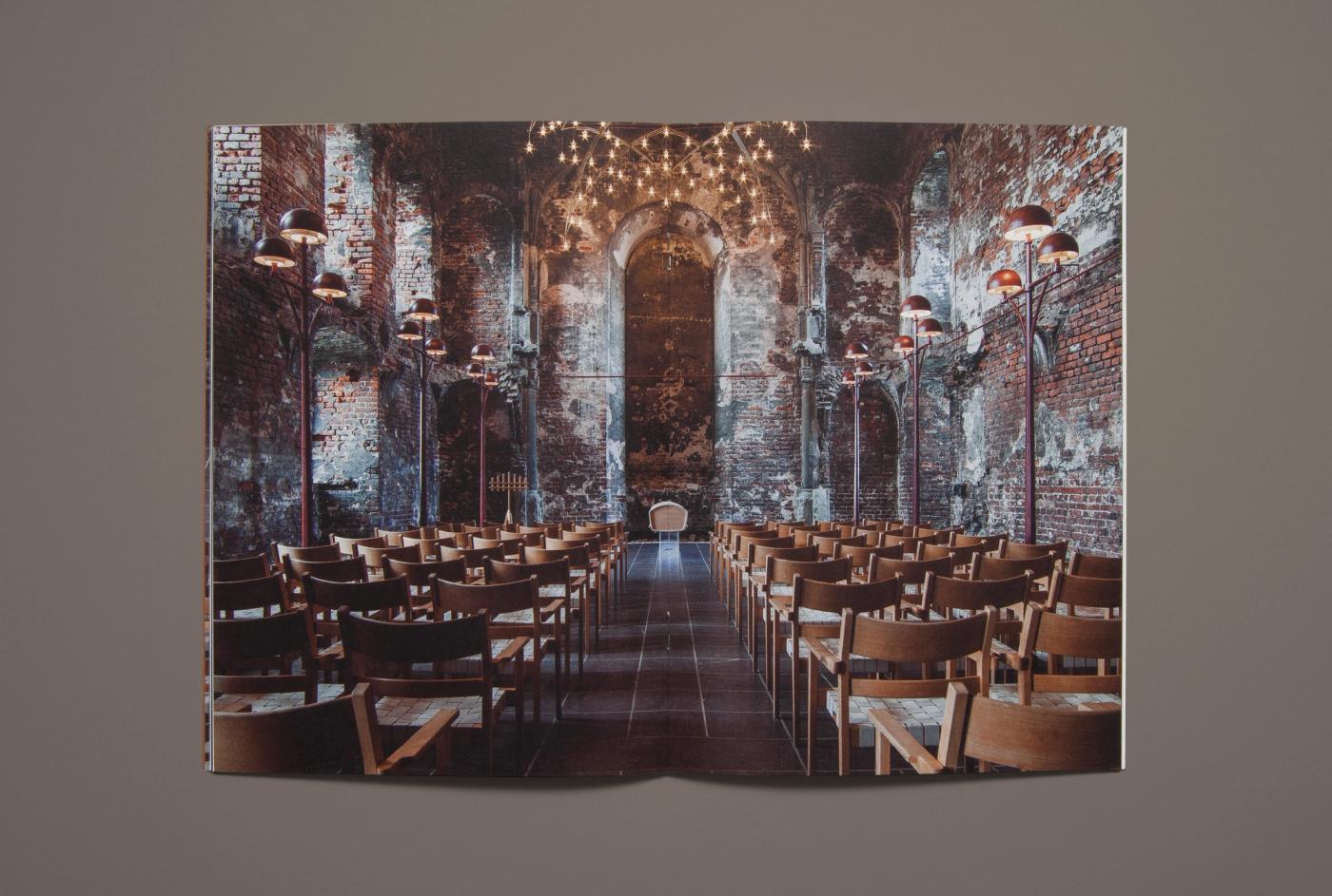 Double-page photograph from the Ro catalogue of a brick hall with rows of wooden chairs, warm lamps along the walls and a chandelier overhead, with a white Akari urn at the far end.