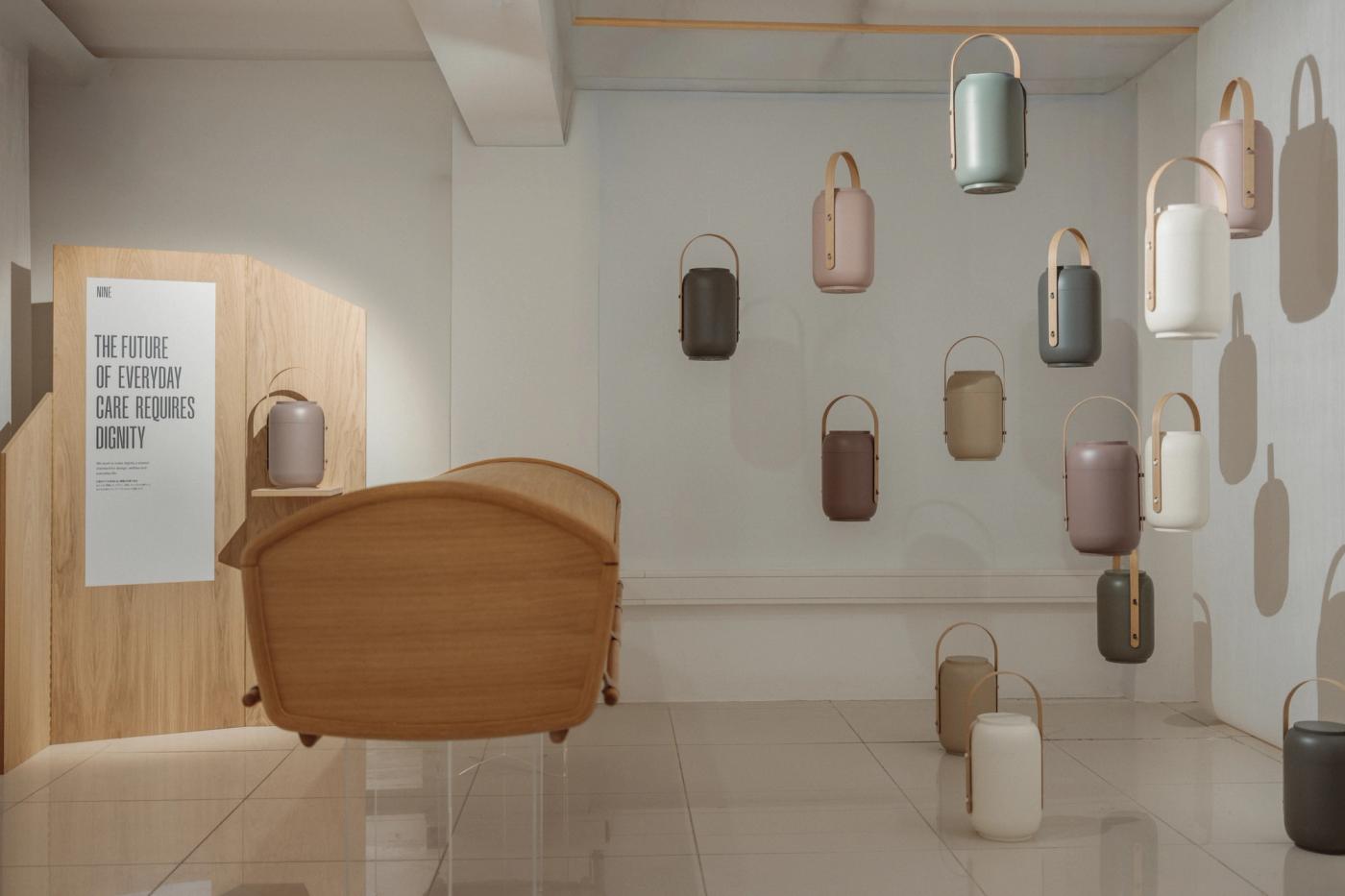 Akari urns in sage, rose, taupe and cream with wooden handles, hanging on a white wall and standing on the floor, beside an oak armchair and an oak panel with the poster "The future of everyday care requires dignity".