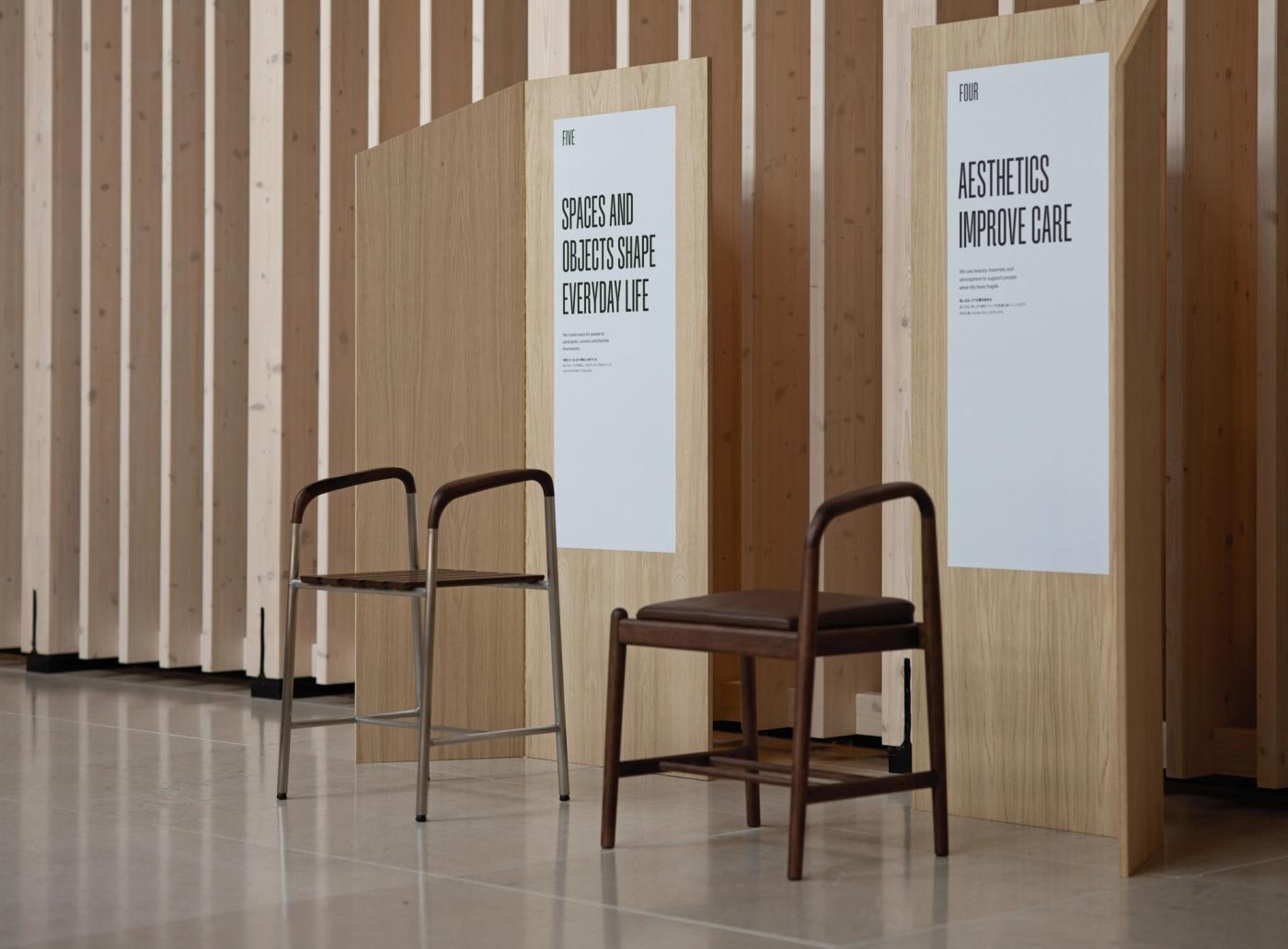 Bath stool in stainless steel and wood beside a stool in dark-stained wood with a brown leather seat, placed in front of wooden panels with the posters "Spaces and Objects Shape Everyday Life" and "Aesthetics Improve Care".