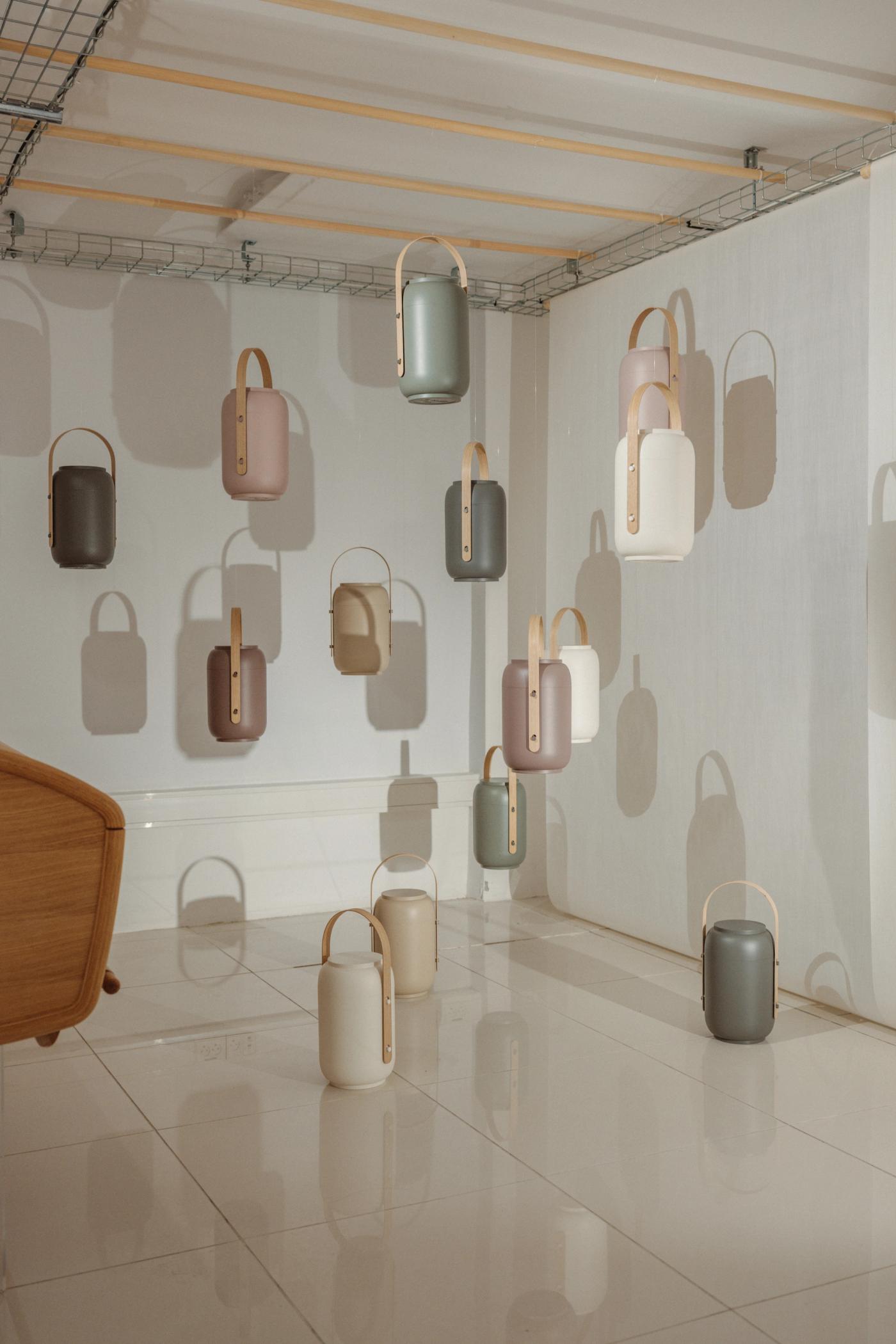 Akari urns in sage, rose, cream, taupe and grey with wooden handles, hanging on a white wall and standing on a tiled floor, beside an oak armchair.