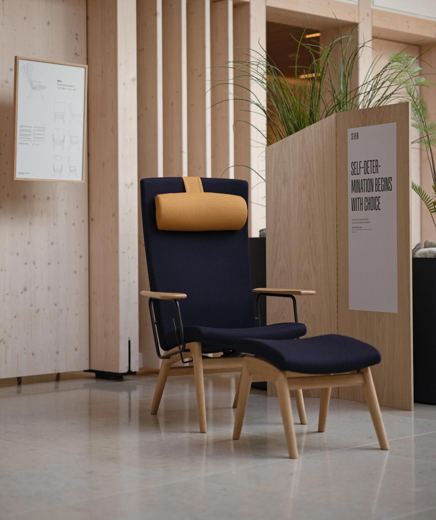HIRO lounge chair with an ochre headrest and light wooden legs and armrests, placed in a bright interior beside a wooden panel with a poster and a tall plant.