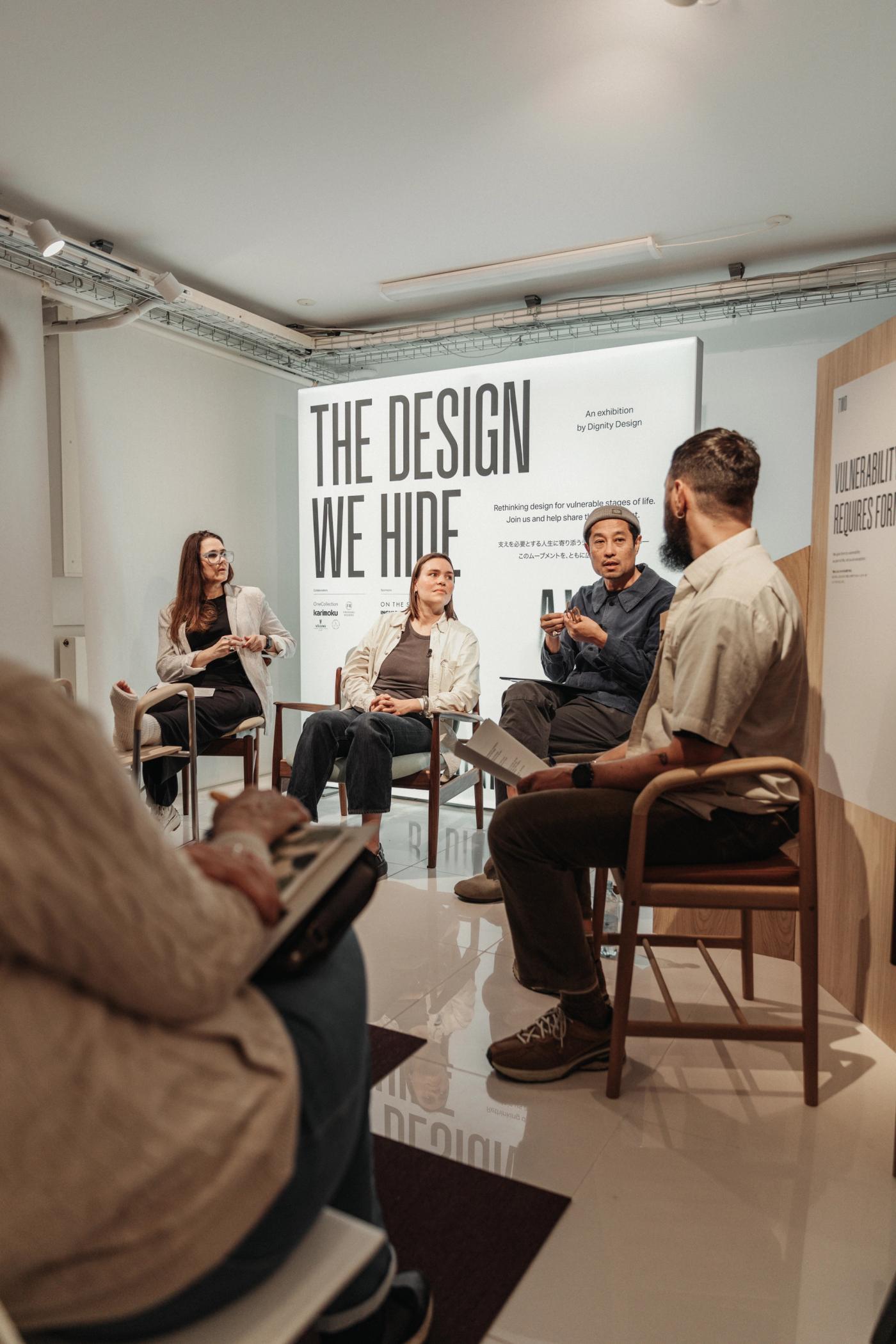 Panel discussion at The Design We Hide Away with four speakers seated on wooden chairs in front of a projection of the exhibition title, with an audience member in the foreground.