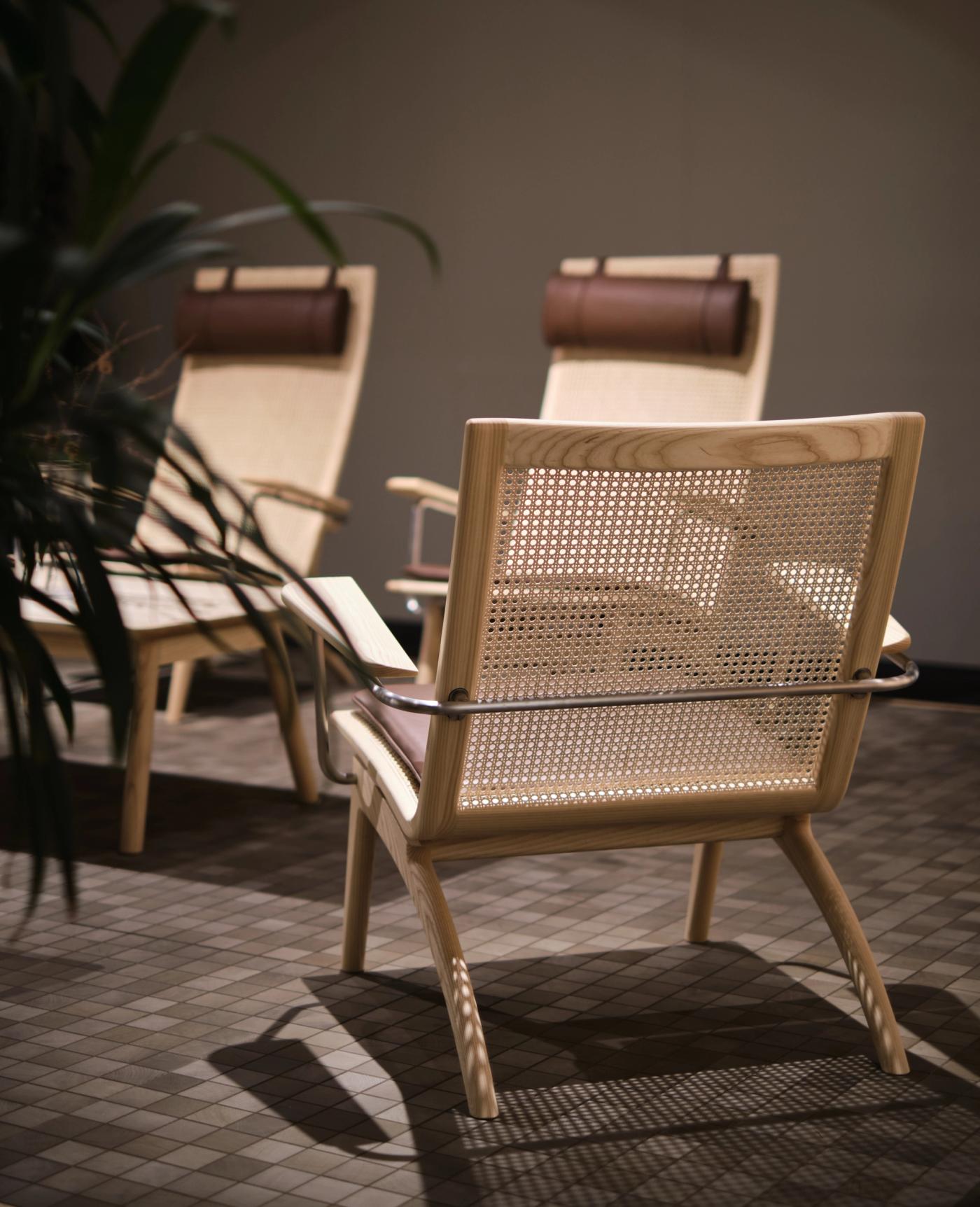 GAZE lounge chair with a woven rattan back and a steel armrest, seen from behind, with two GAZE high-back chairs with leather headrests in the background beside a tall plant.