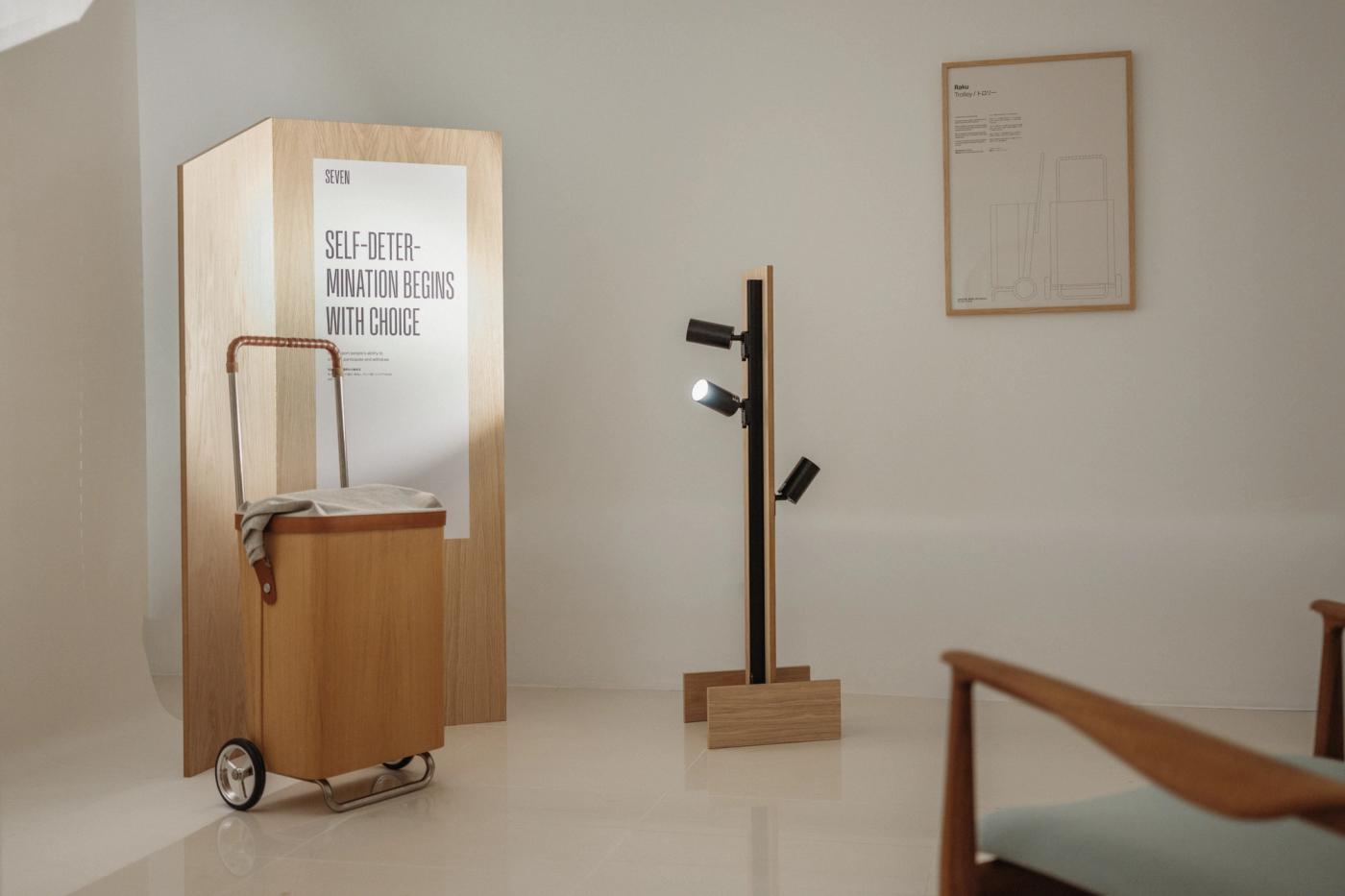 Raku trolley in oak with a leather-wrapped handle beside an oak panel with the poster "Self-determination begins with choice", with a black and oak lamp and a framed drawing on the white wall behind.