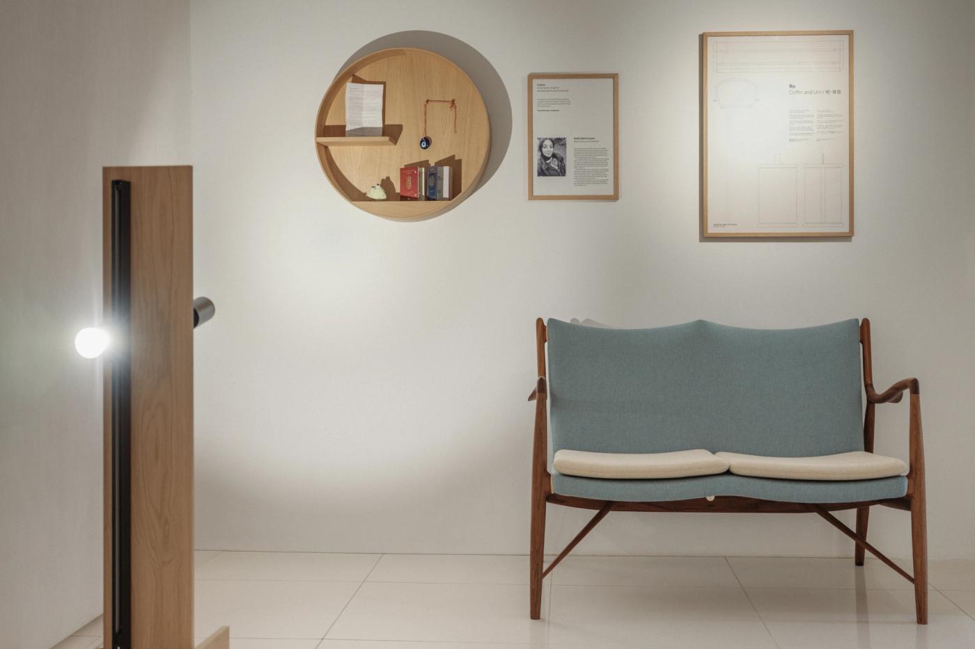 Round Cyklus memorial shelf in oak on a white wall, with framed text panels beside it and a wooden two-seat sofa with a mint upholstered back and cream seat cushions in front.
