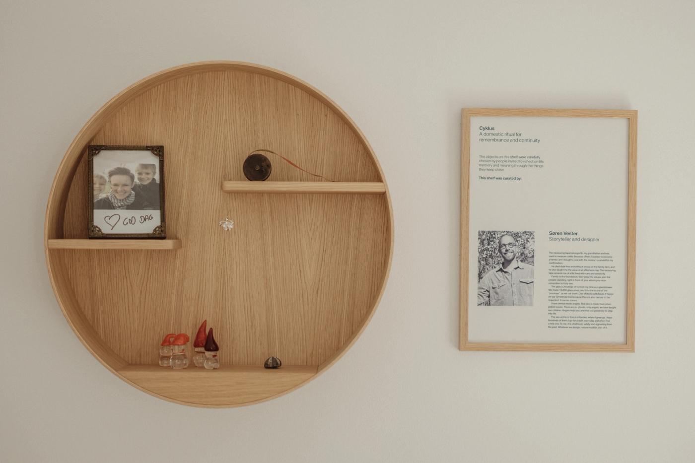 Cyklus memorial shelf in oak, a round wall shelf holding a framed photograph and small objects, beside a framed text panel and a black and oak lamp with two spotlights.