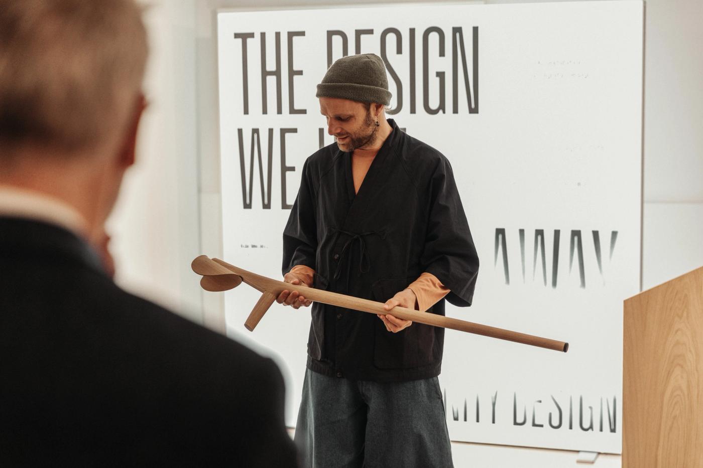 A man in a dark wrap jacket and a grey beanie holds a wooden cane with a curved handle during a talk, in front of a poster reading "The Design We Hide Away by Dignity Design".