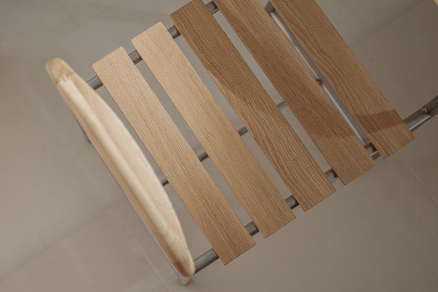 Close-up of the slatted oak seat and curved oak armrest of a bath stool with a stainless steel frame, showing the wood grain and metal beneath.