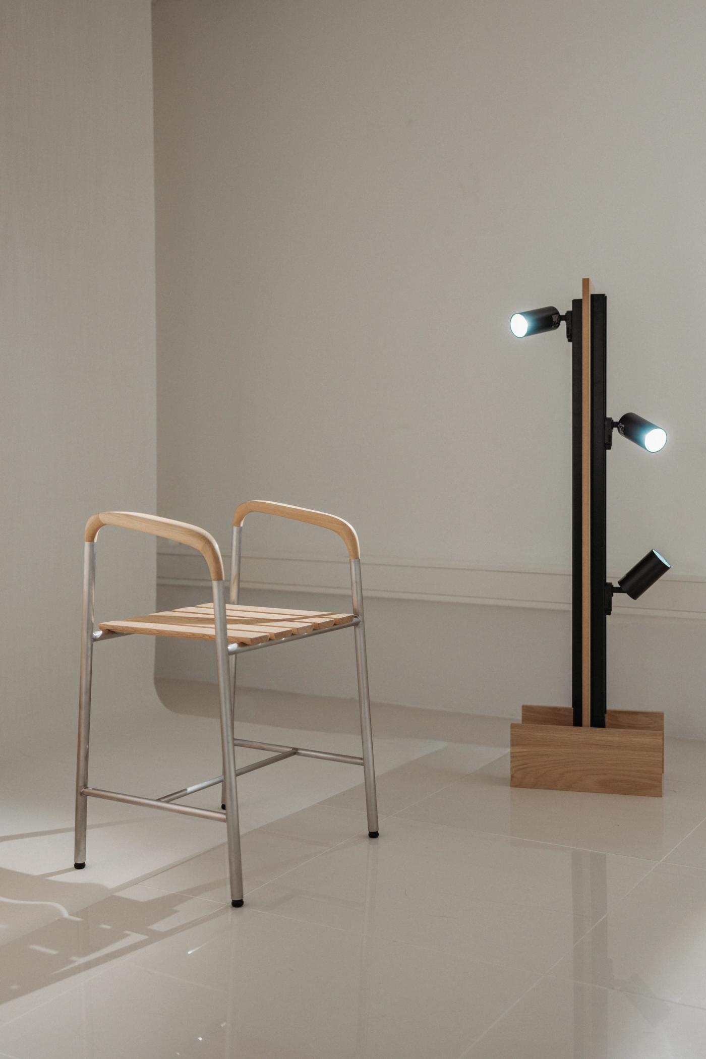 Steel-framed chair with oak armrests and a slatted oak seat beside a floor lamp with three adjustable spotlights on a black and oak column, on a glossy white floor.  Jeg kunne ikke finde et produkt på hjemmesiden, der matcher denne stol eller lampe, så beskrivelsen er generel.