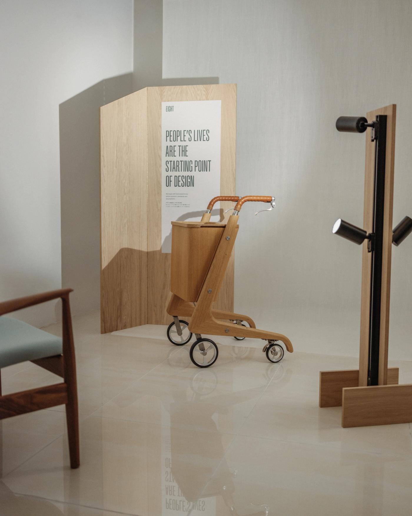 Sindig wooden walker with a leather-wrapped handle and four wheels in front of an oak panel with the poster "People's lives are the starting point of design", beside a black and oak lamp.