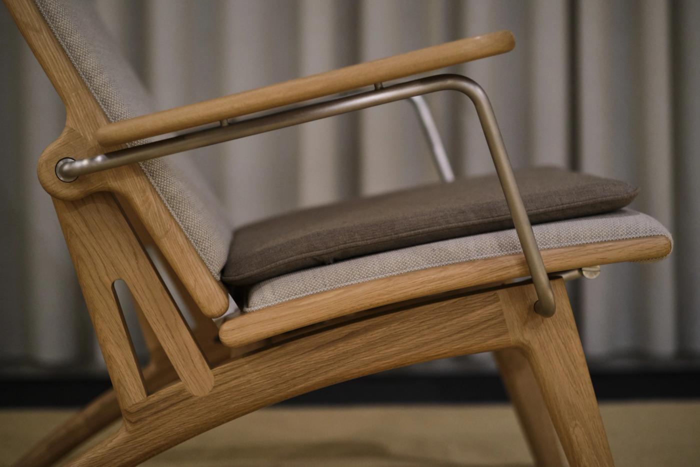 Detail of a GAZE chair's oak frame with a curved oak armrest, a steel support and light and grey upholstery against grey curtains.