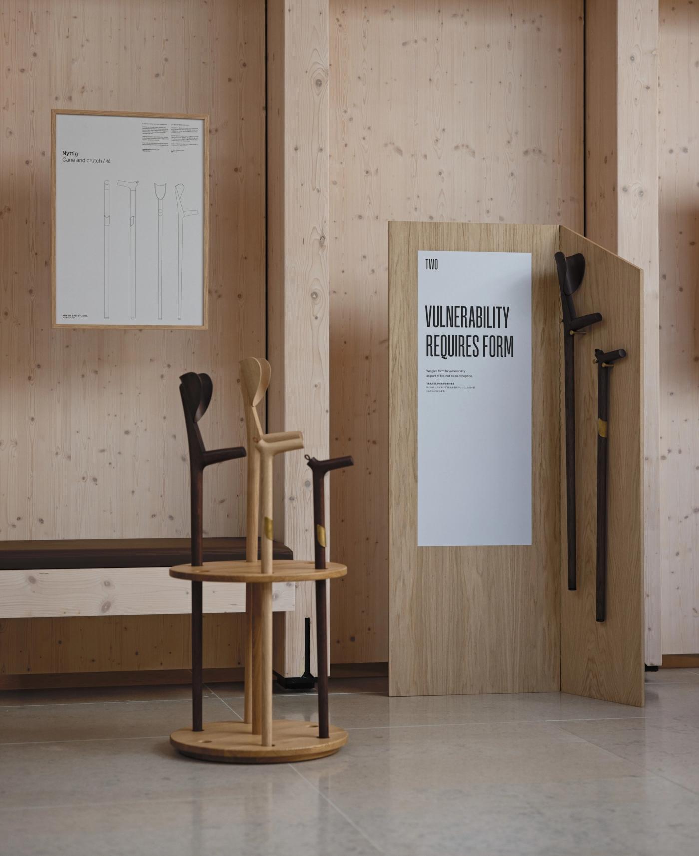 Wooden walking sticks and crutches in dark and light wood, standing on a round wooden stand beside a wooden panel with the poster "Vulnerability Requires Form".