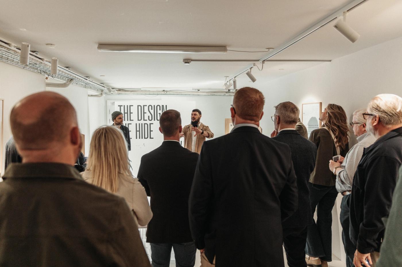 Visitors gathered in a white gallery listening to a speaker in front of a wall with the exhibition title "The Design We Hide Away".