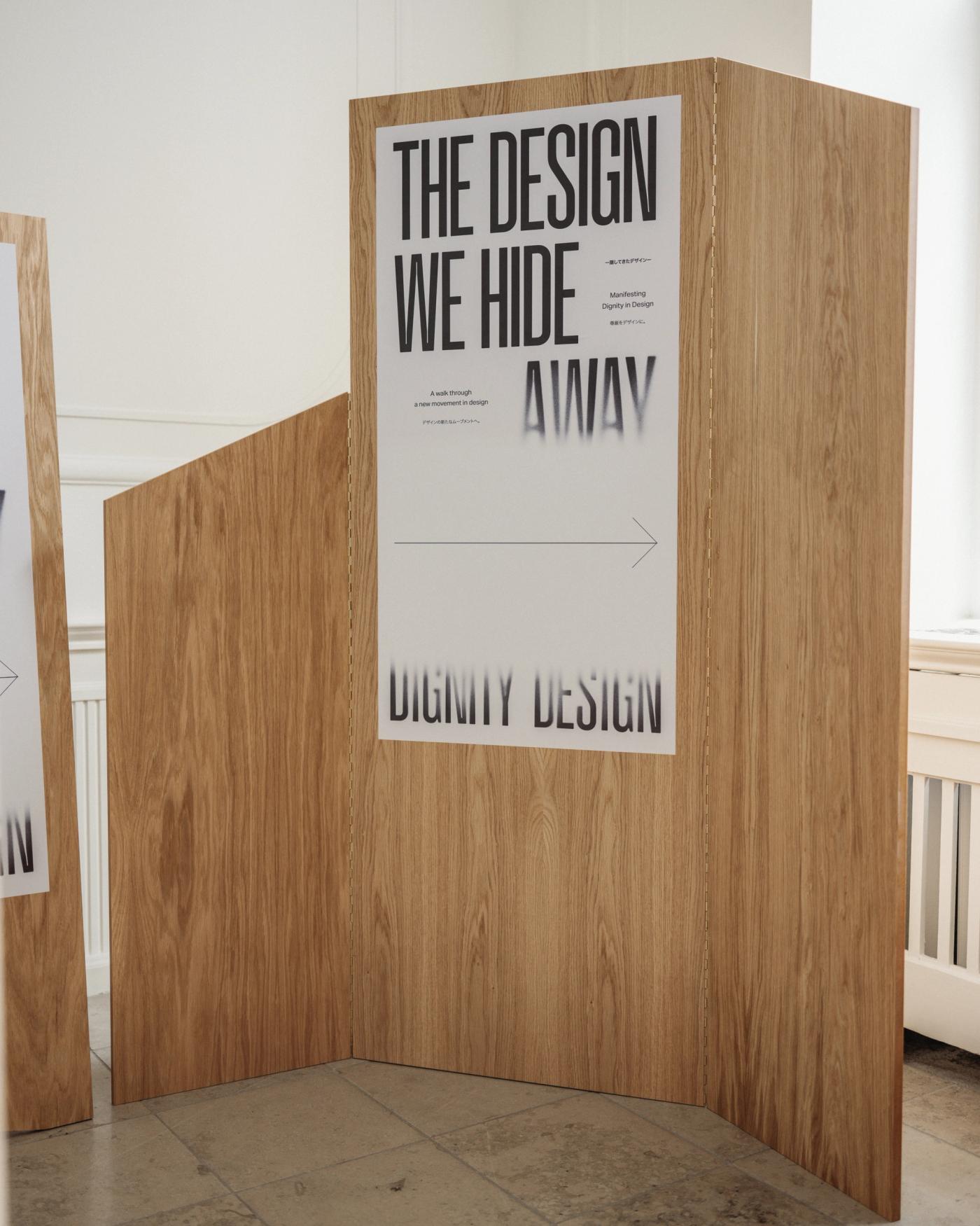 Oak wall panel with the poster "The Design We Hide Away by Dignity Design" and an arrow pointing right, on a stone floor beside a white wall with a radiator.